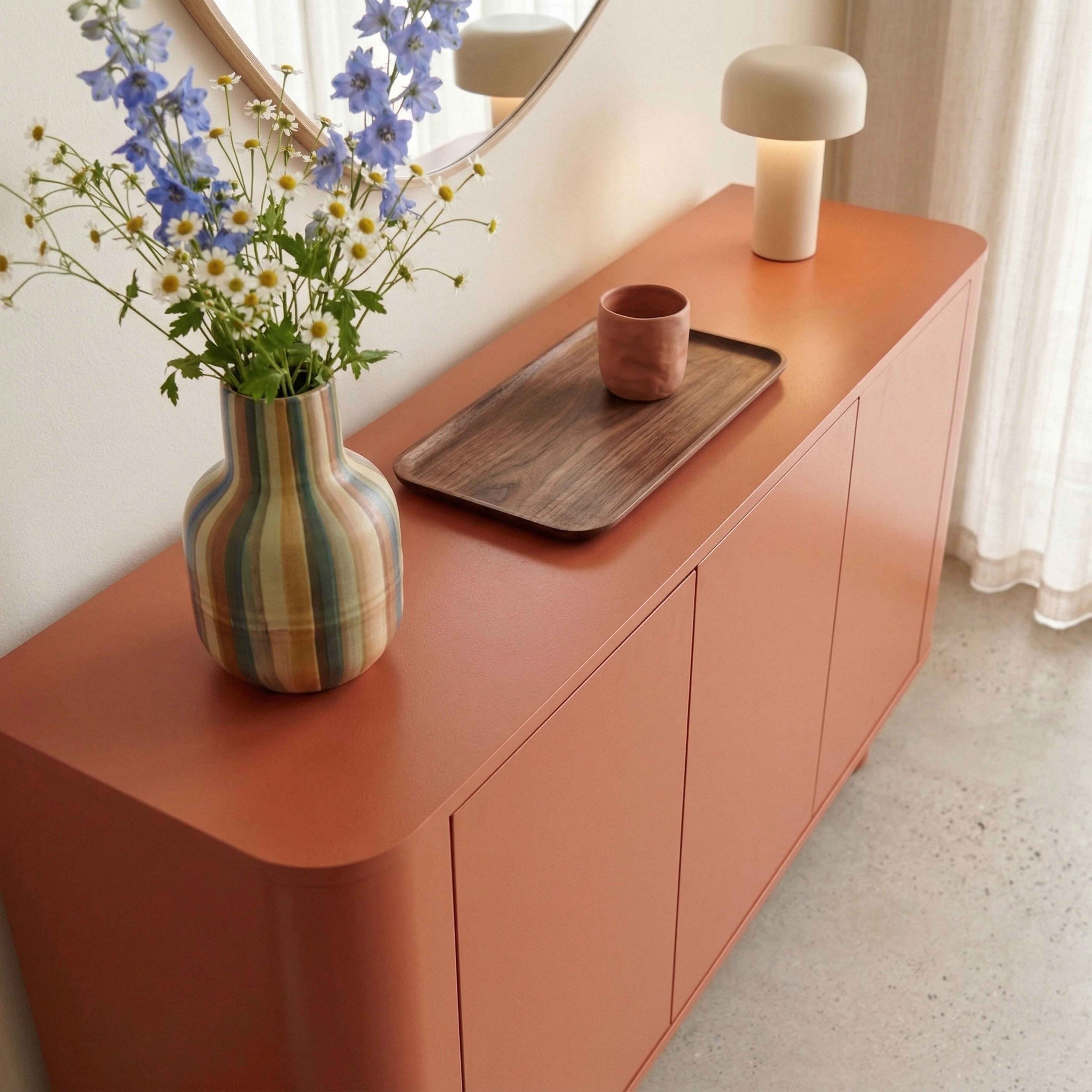 Ein korallenfarbenes Sideboard mit Blumenstrauß, Tablett, Tasse und Lampe.