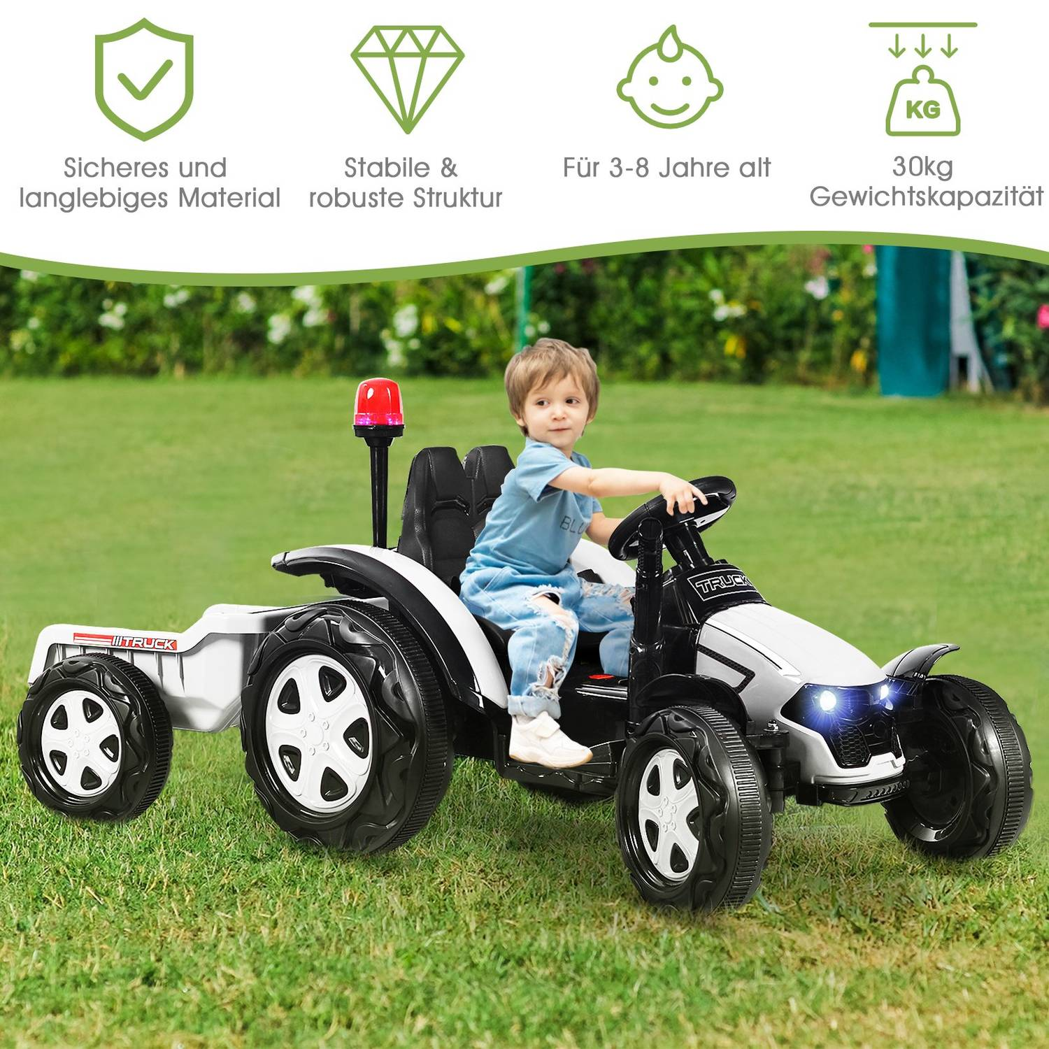 A child rides a white and black toy tractor with a trailer on grass.