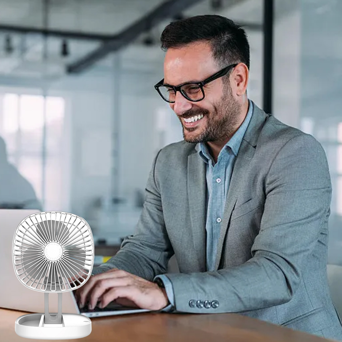 Mann im Anzug lächelt und tippt auf einem Laptop, kleiner Ventilator daneben.