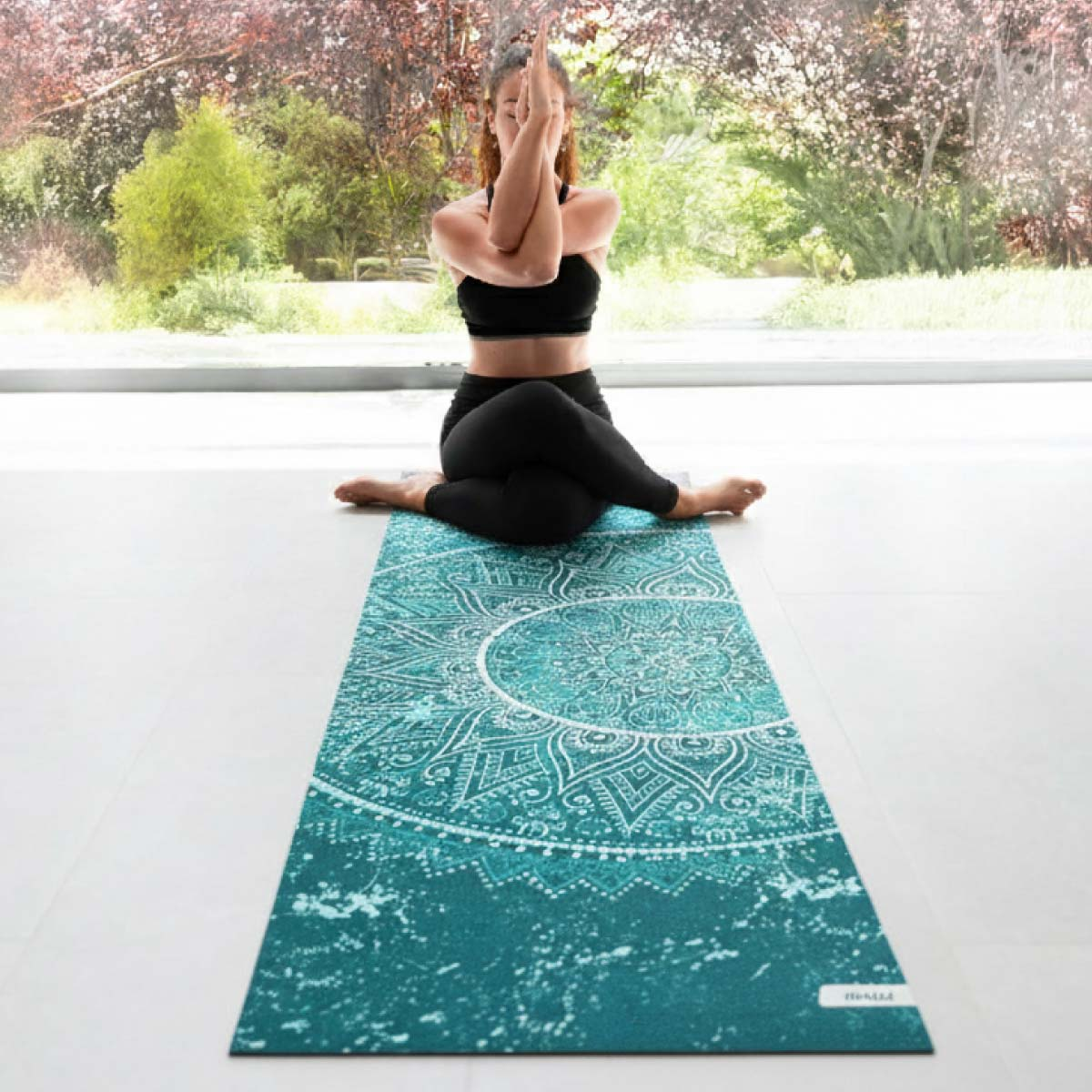 Mujer con ropa deportiva negra practica yoga sobre una esterilla turquesa con mandala.
