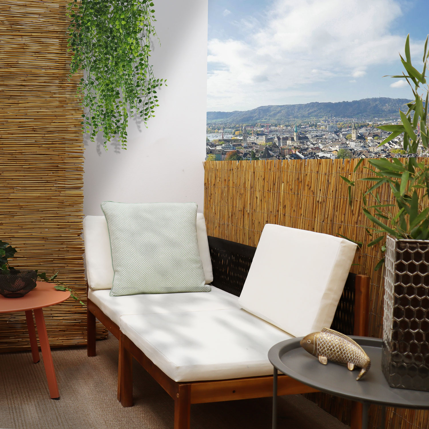 Eine Liege mit weißen Kissen auf einem Balkon mit Stadtblick. Bambusmatten im Hintergrund.