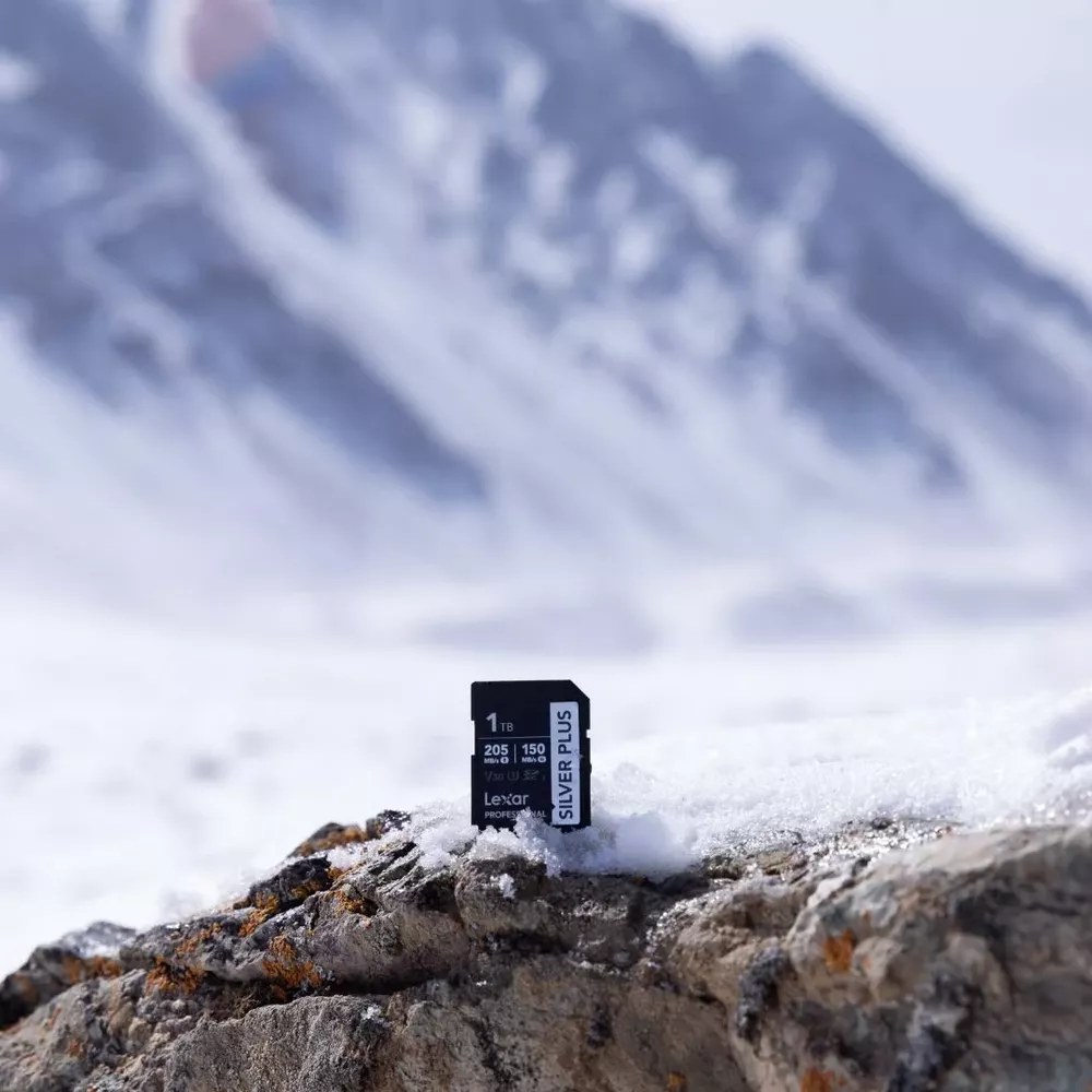 Una scheda SD Lexar da 1TB riposa su una roccia nella neve, montagne sullo sfondo.