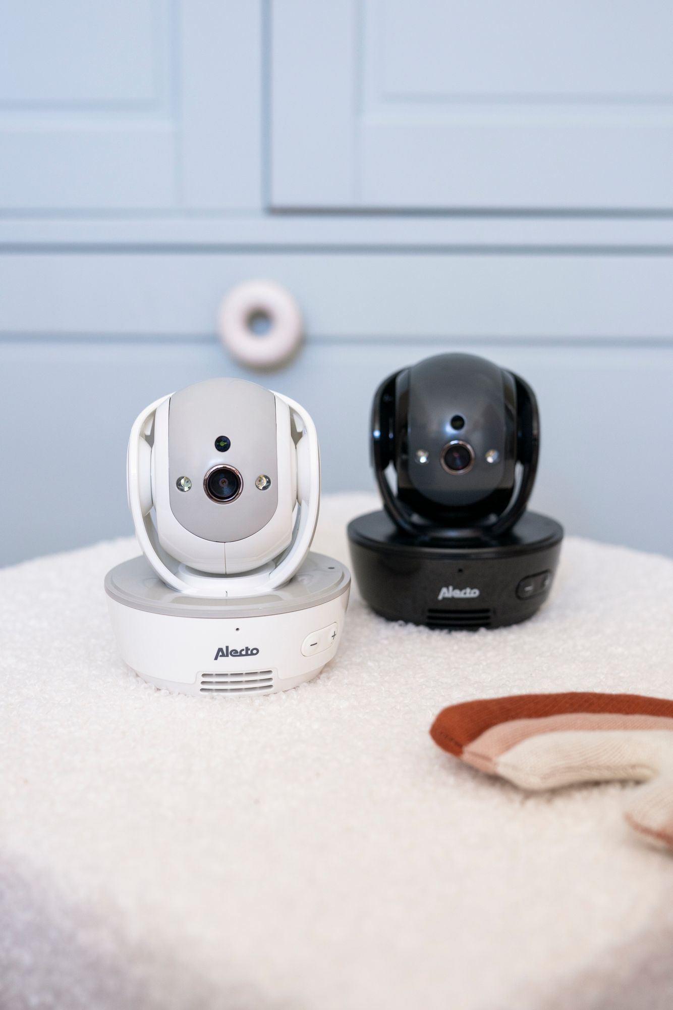 Zwei Babyphones, weiß und schwarz, auf einem weißen Teppich vor einer blauen Wand.