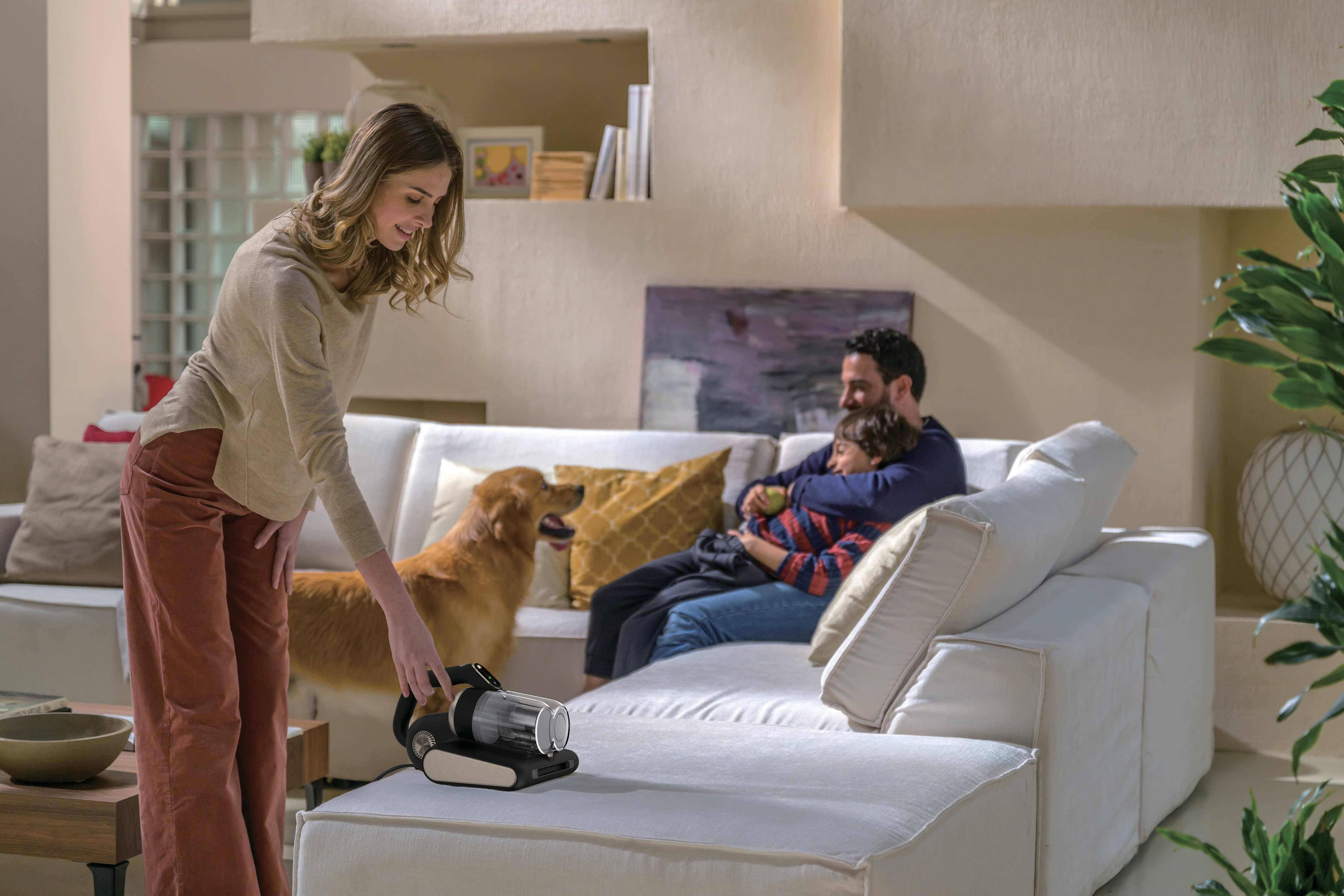 Donna con aspirapolvere su divano, famiglia e cane sullo sfondo.