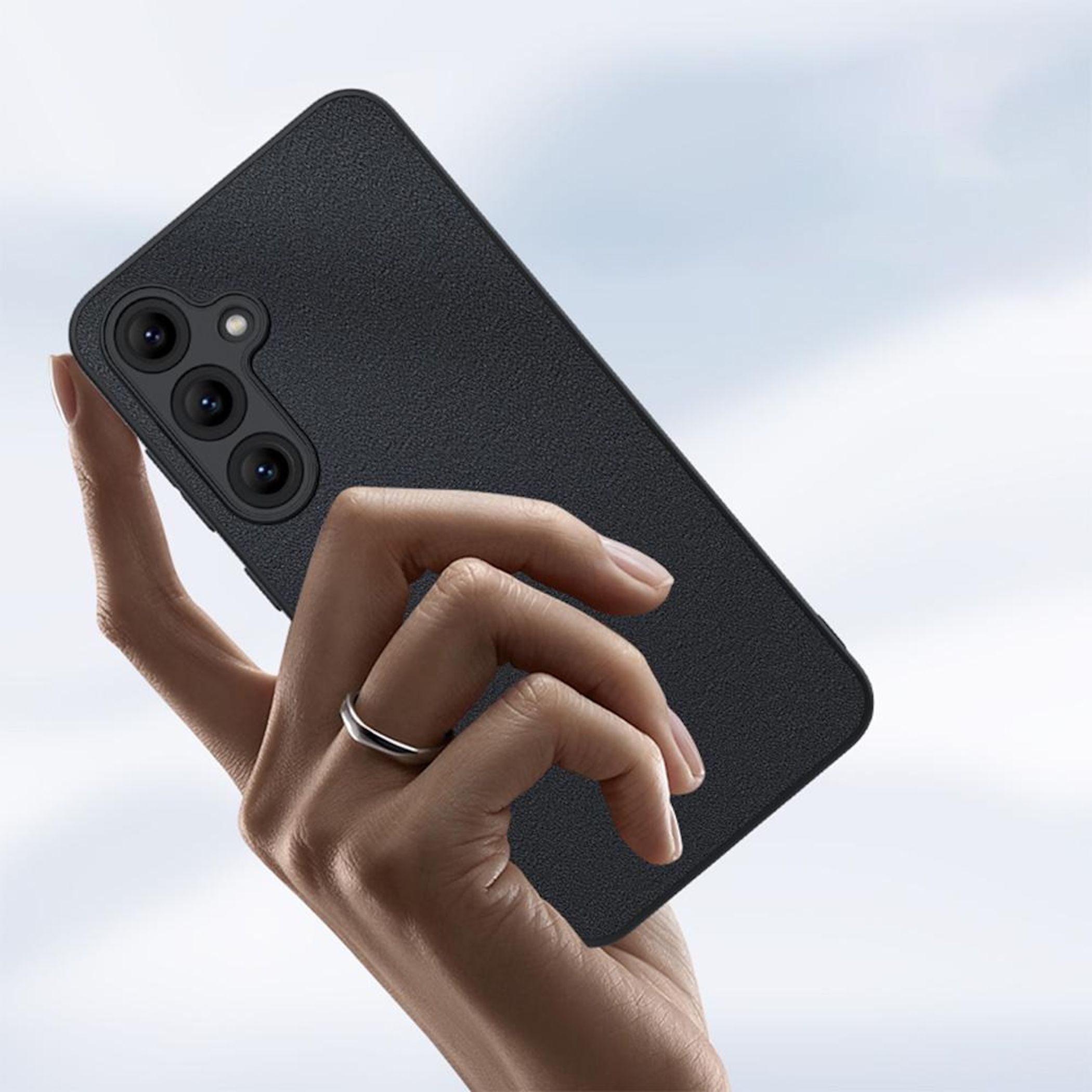 Una mano sostiene un teléfono negro con tres cámaras. Un anillo en el dedo.