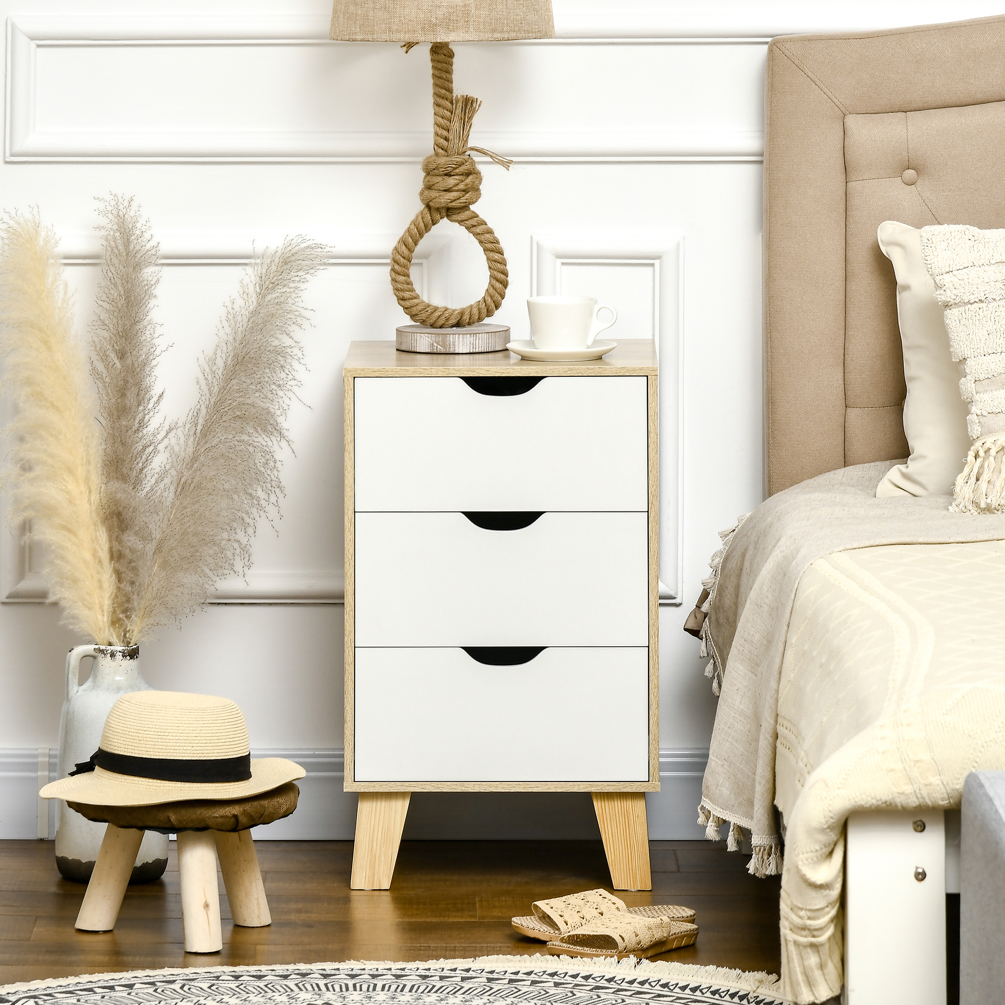 Una mesita de noche con cajones blancos junto a una cama. Un sombrero de paja descansa sobre un taburete de madera.