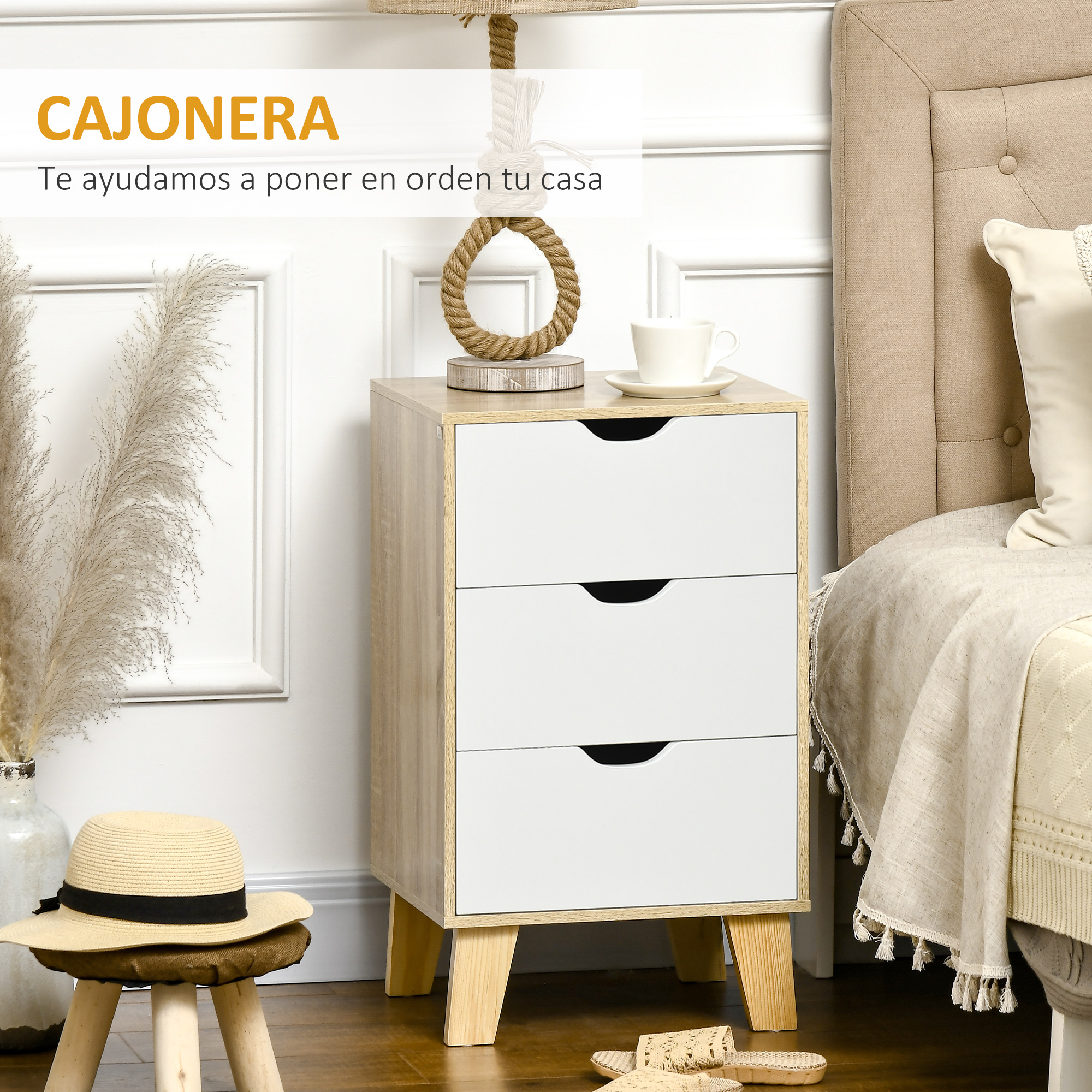 Una mesita de noche de tres cajones de madera clara y blanca junto a una cama, con un sombrero de paja y hierba de la Pampa cerca.