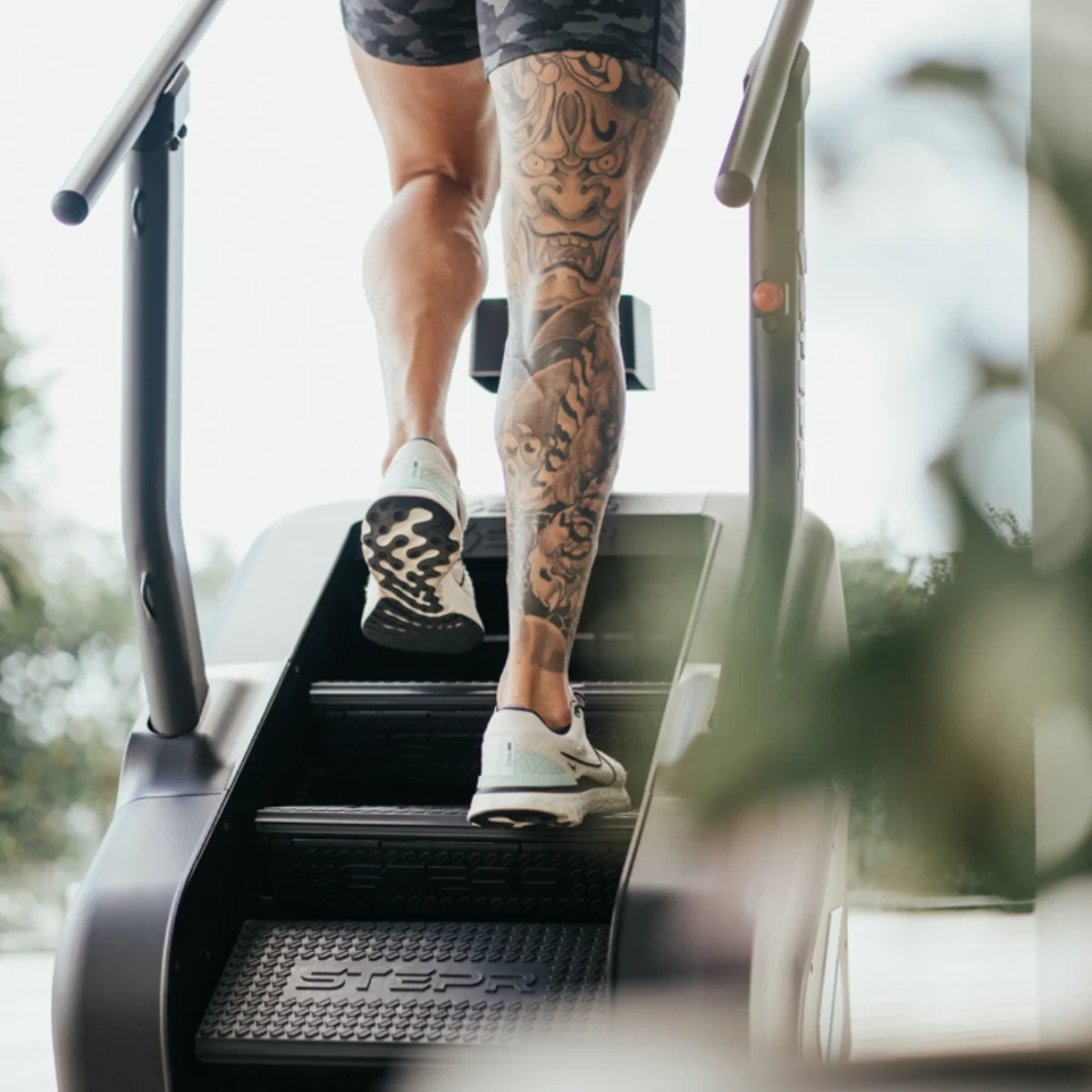Mann mit Tattoos steigt eine Treppenmaschine hinauf, trägt Turnschuhe.