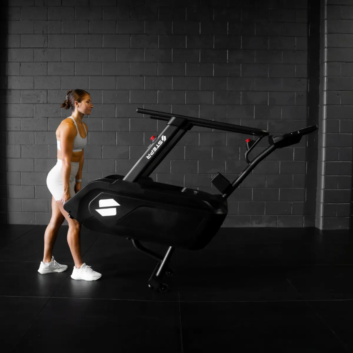 Frau in weißer Sportkleidung steht neben einem schwarzen Laufband im Fitnessstudio.