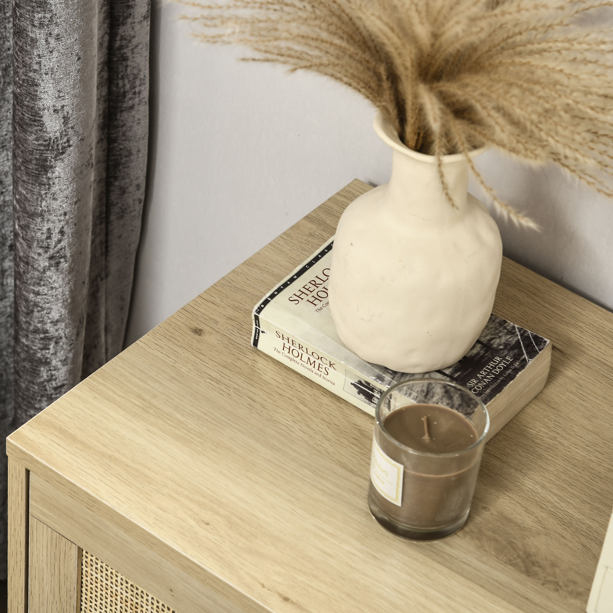 Mesita de noche de madera con libro, vela y jarrón de flores secas. Cortina gris.