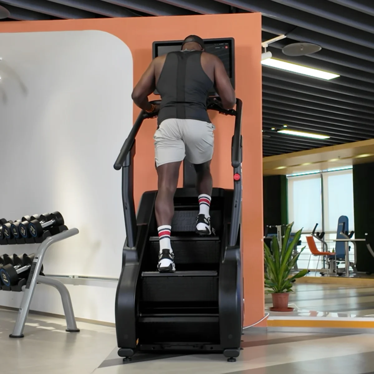 A man exercises on a stair climber in a gym, with dumbbells in the foreground.