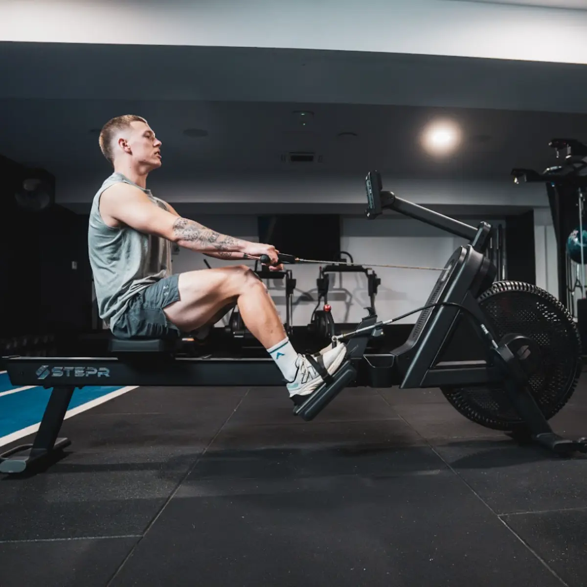 Man roeit op een machine in een sportschool, draagt een grijs tanktop en een korte donkere broek.
