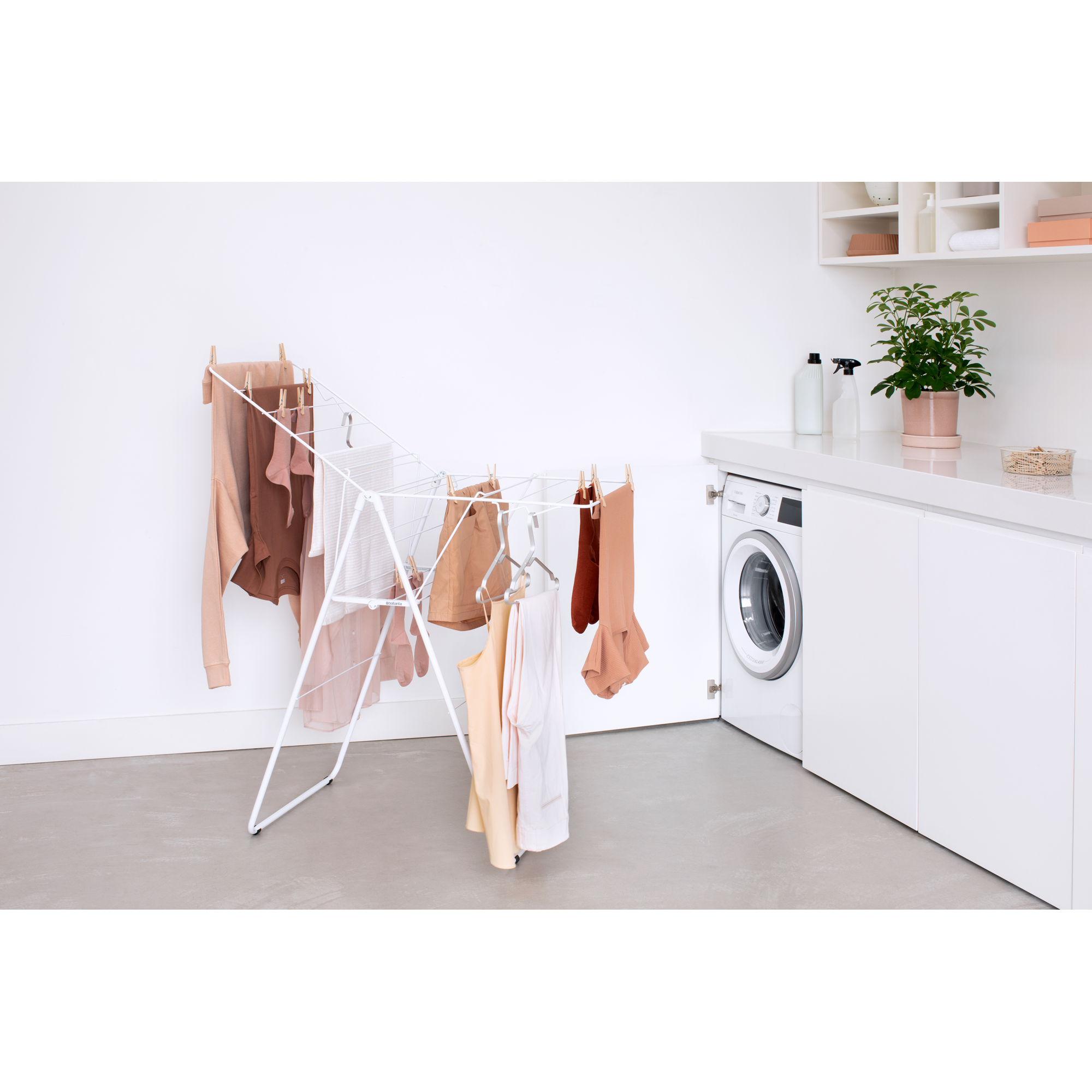 A white clothes drying rack with beige and brown clothes, next to a washing machine.
