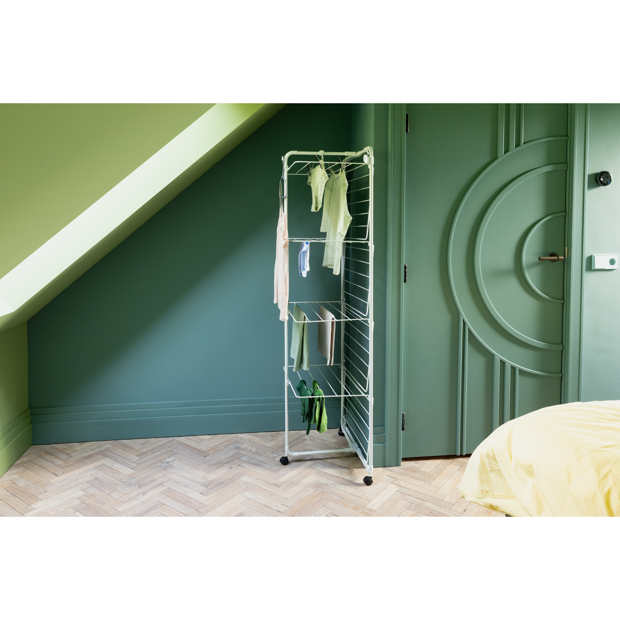 Een wit wasrek met kleding staat op een houten vloer tegen een groene muur.