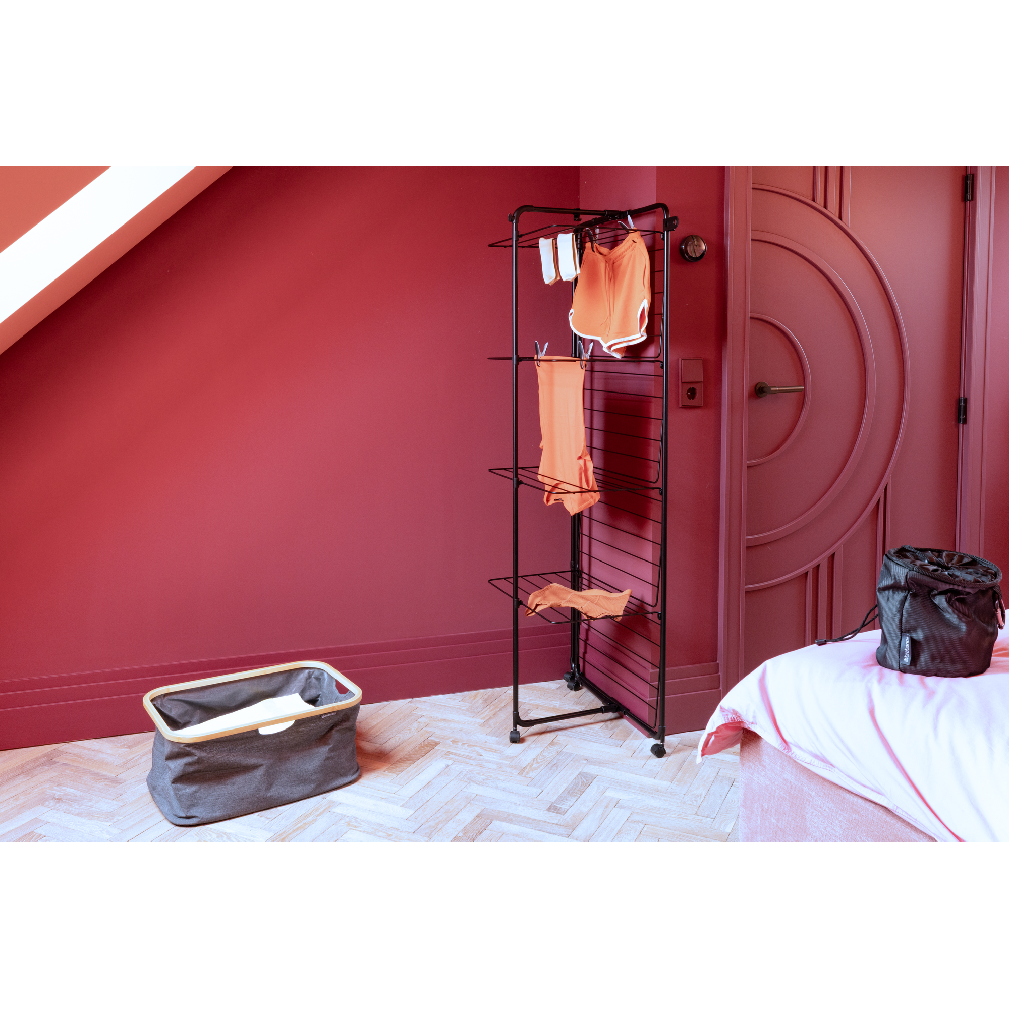 Een droogrek met kleding staat naast een bed en een wasmand.