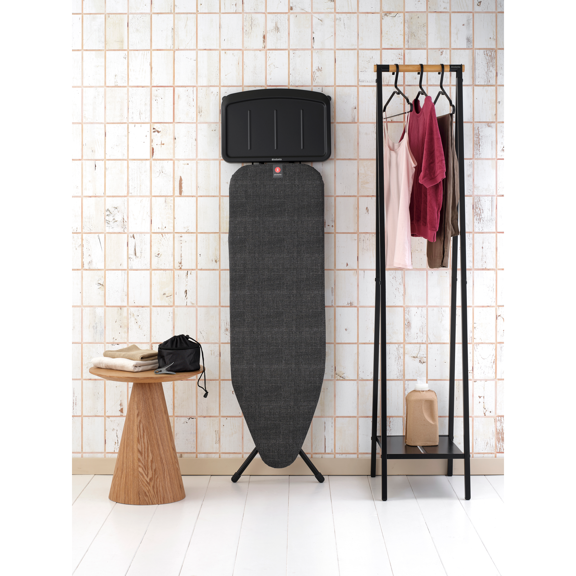 An ironing board stands next to a clothes rack with garments and a wooden table with laundry.