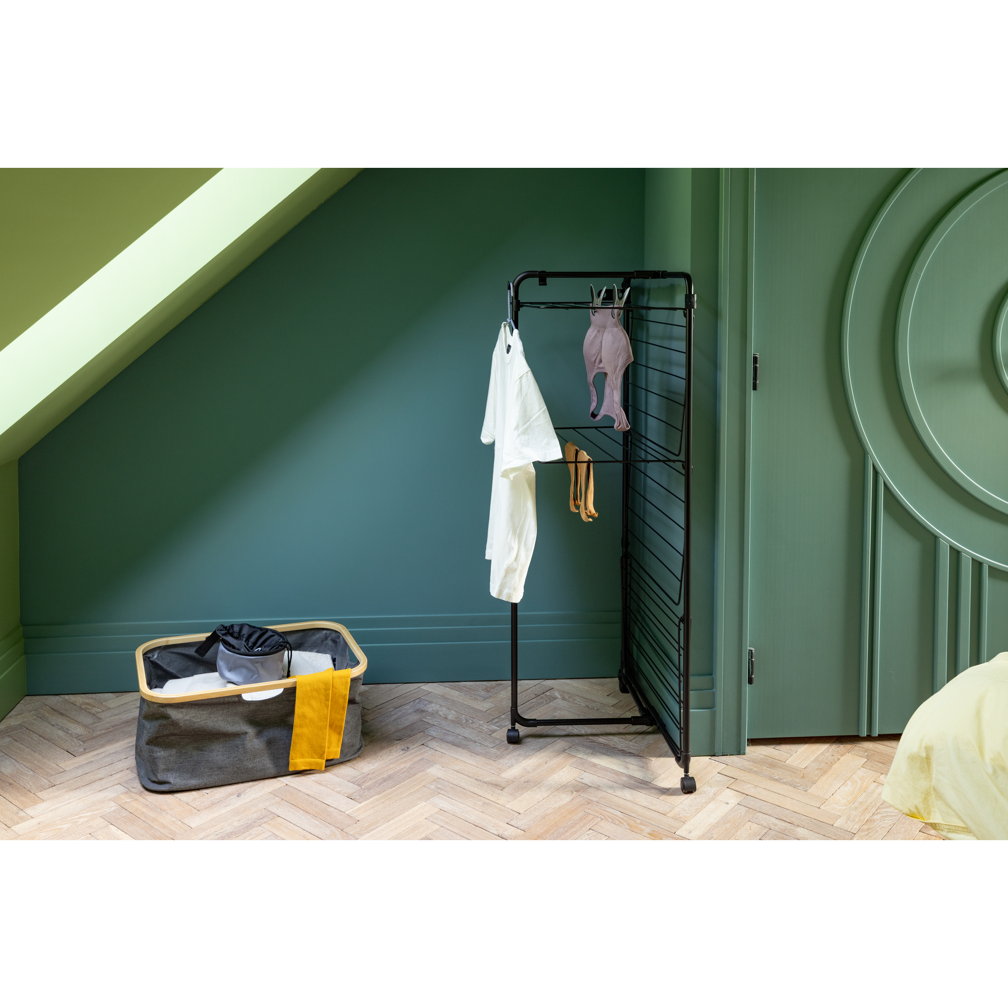 A laundry basket and drying rack stand on a wooden floor in a room with green walls.
