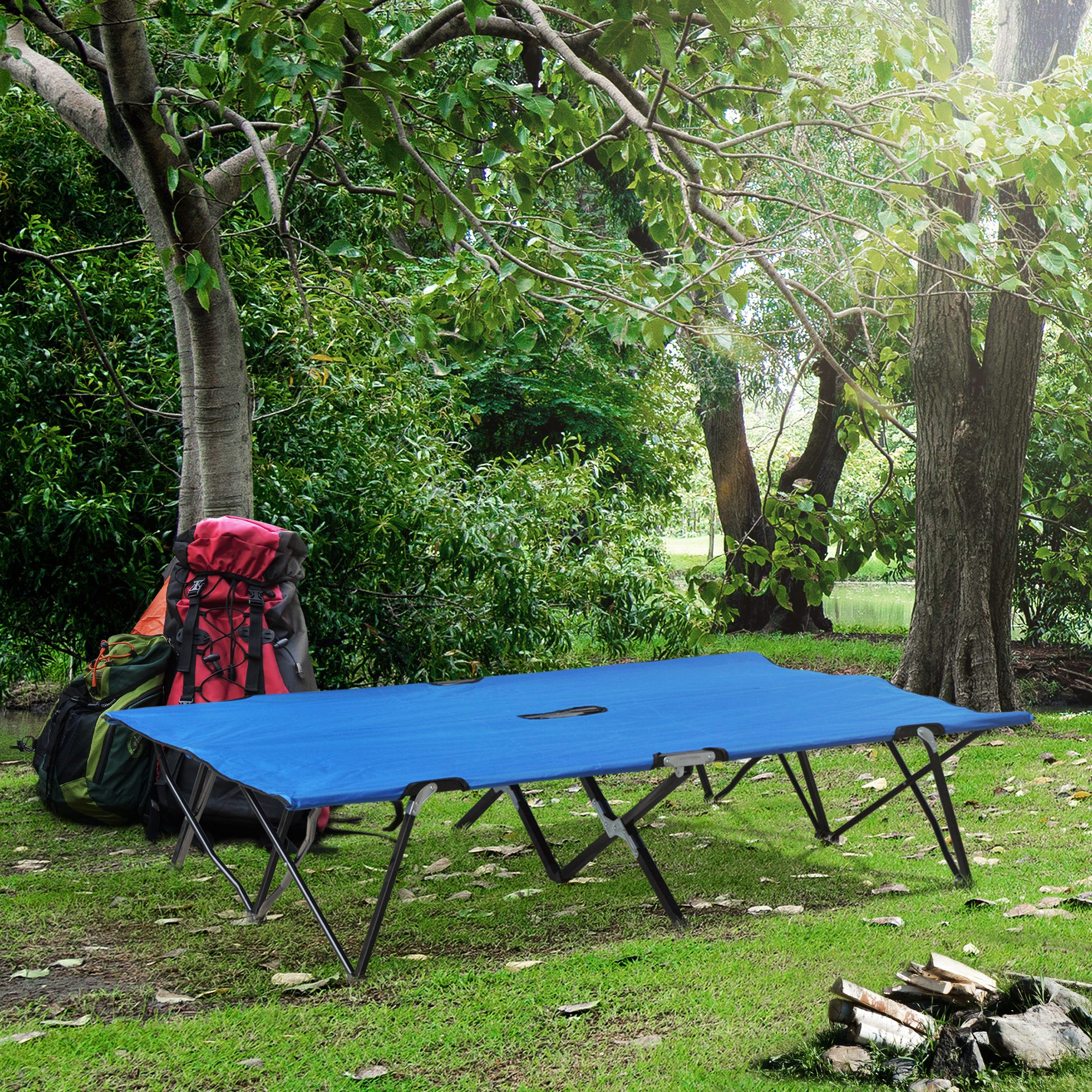 Una cama de camping azul descansa sobre hierba junto a mochilas y leña.