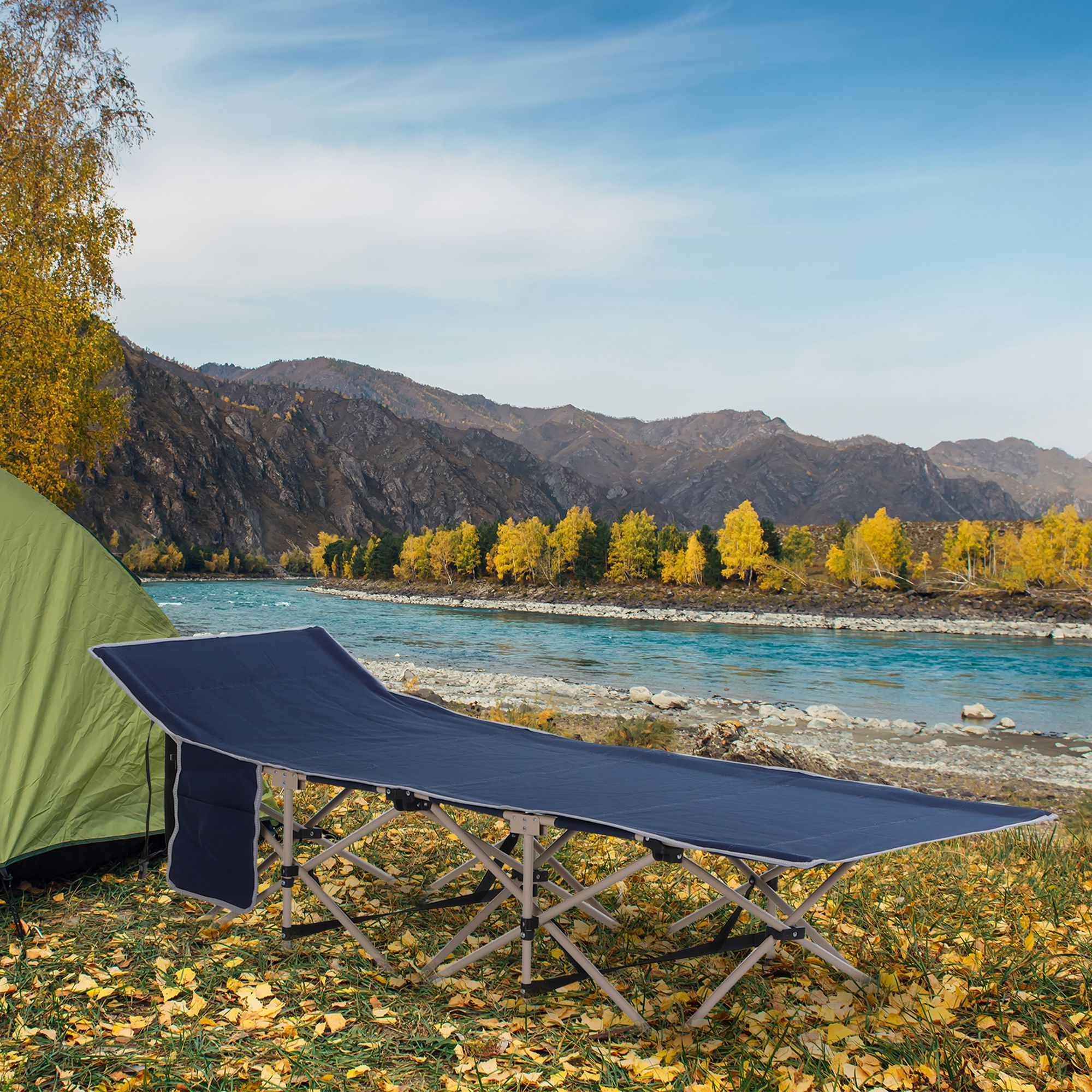 Una tienda verde y una cama plegable azul junto a un río con montañas al fondo.