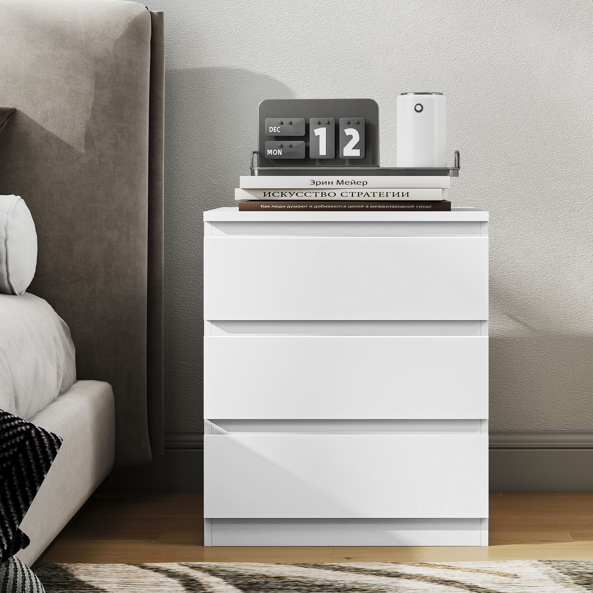 Una mesita de noche blanca de tres cajones junto a una cama. Libros y un calendario encima.