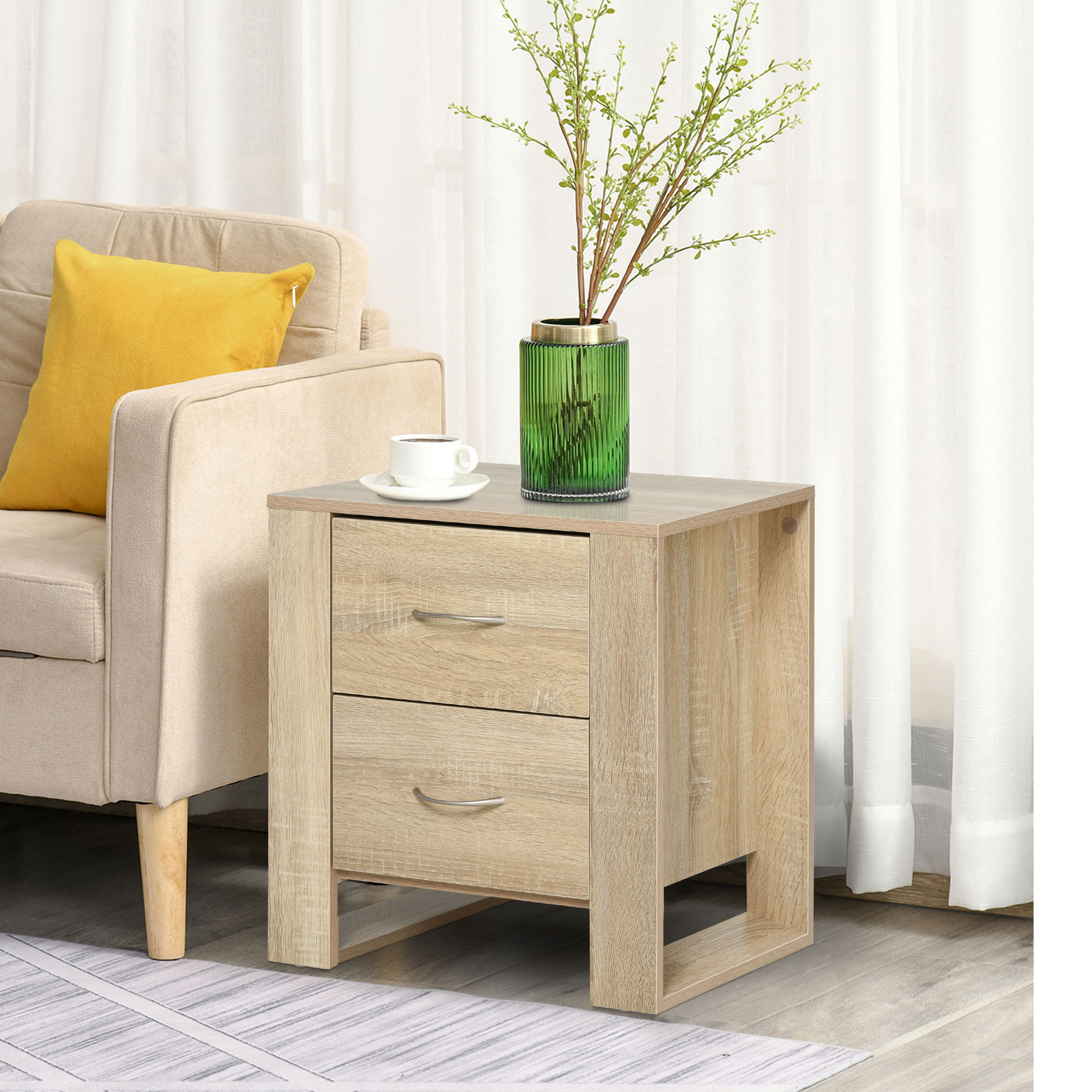 Una mesita de noche de madera con dos cajones junto a un sillón. Un jarrón verde con ramas y una taza de café están encima.
