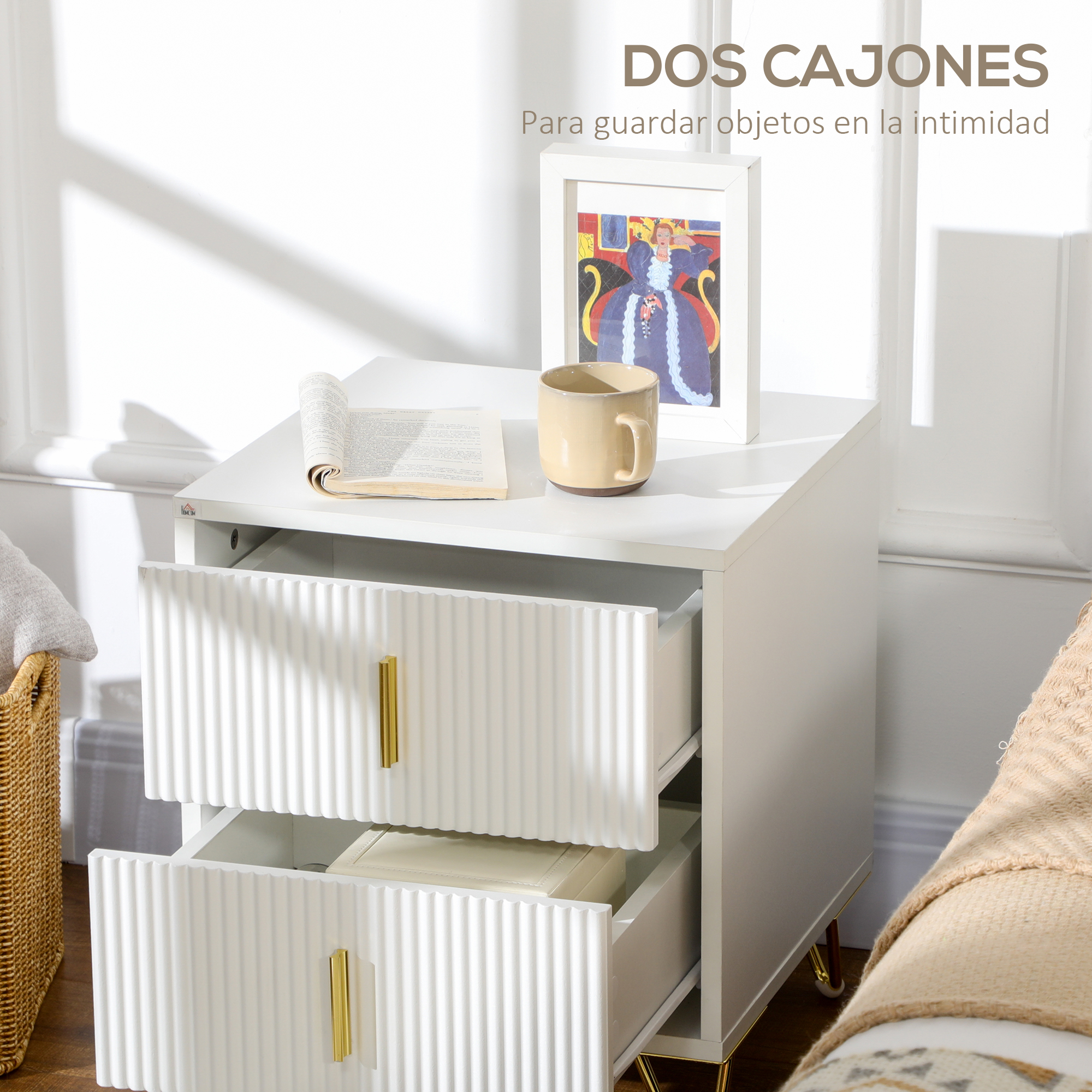Una mesita de noche blanca con cajones estriados y tiradores dorados tiene libro, taza y arte enmarcado.
