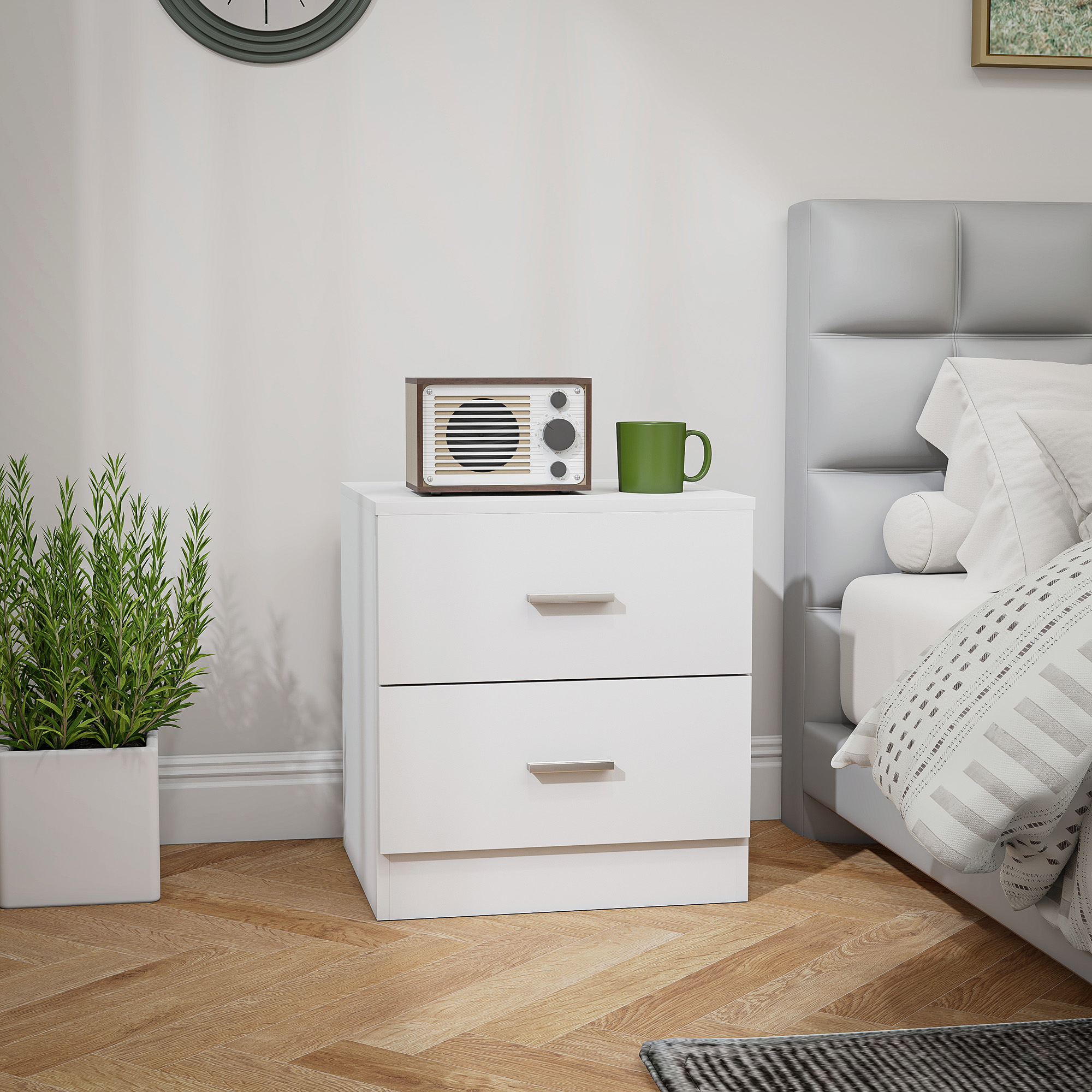 Una mesita de noche blanca con radio y taza verde. Al lado hay una cama.