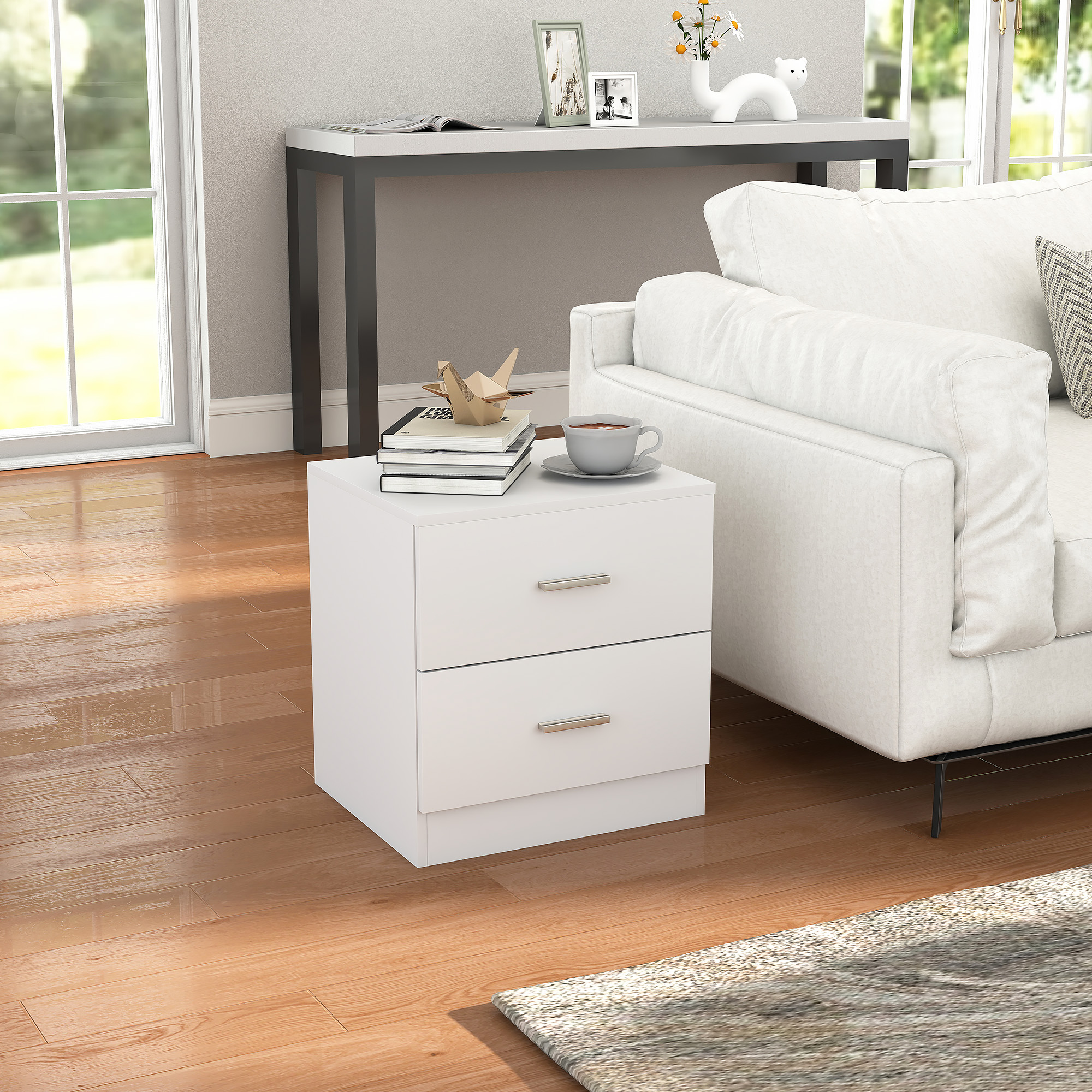 Mesita de noche blanca con dos cajones, junto a un sofá. Encima hay una pila de libros y una taza de café.