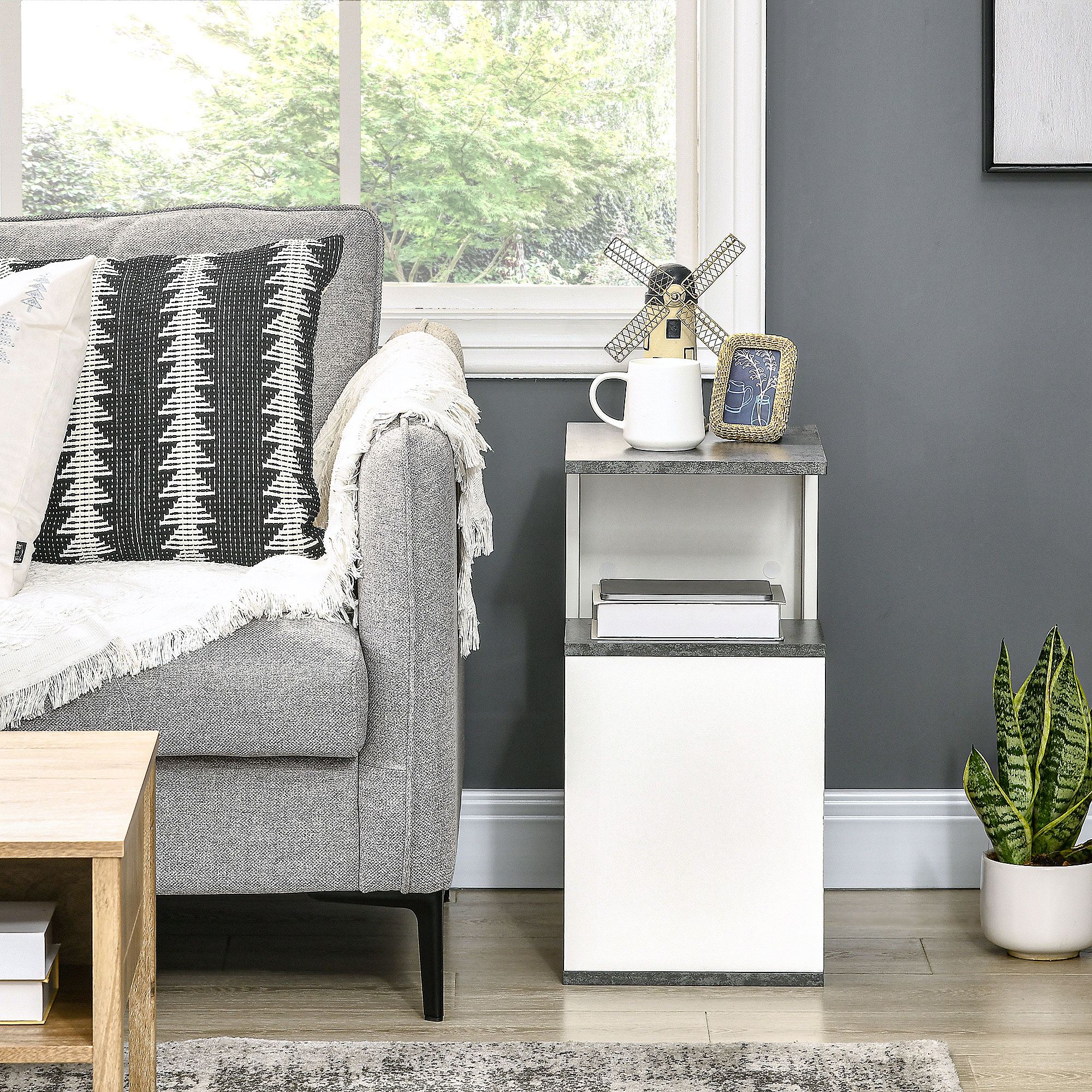 Sofá gris con cojines estampados junto a mesa auxiliar blanca y gris con decoración.