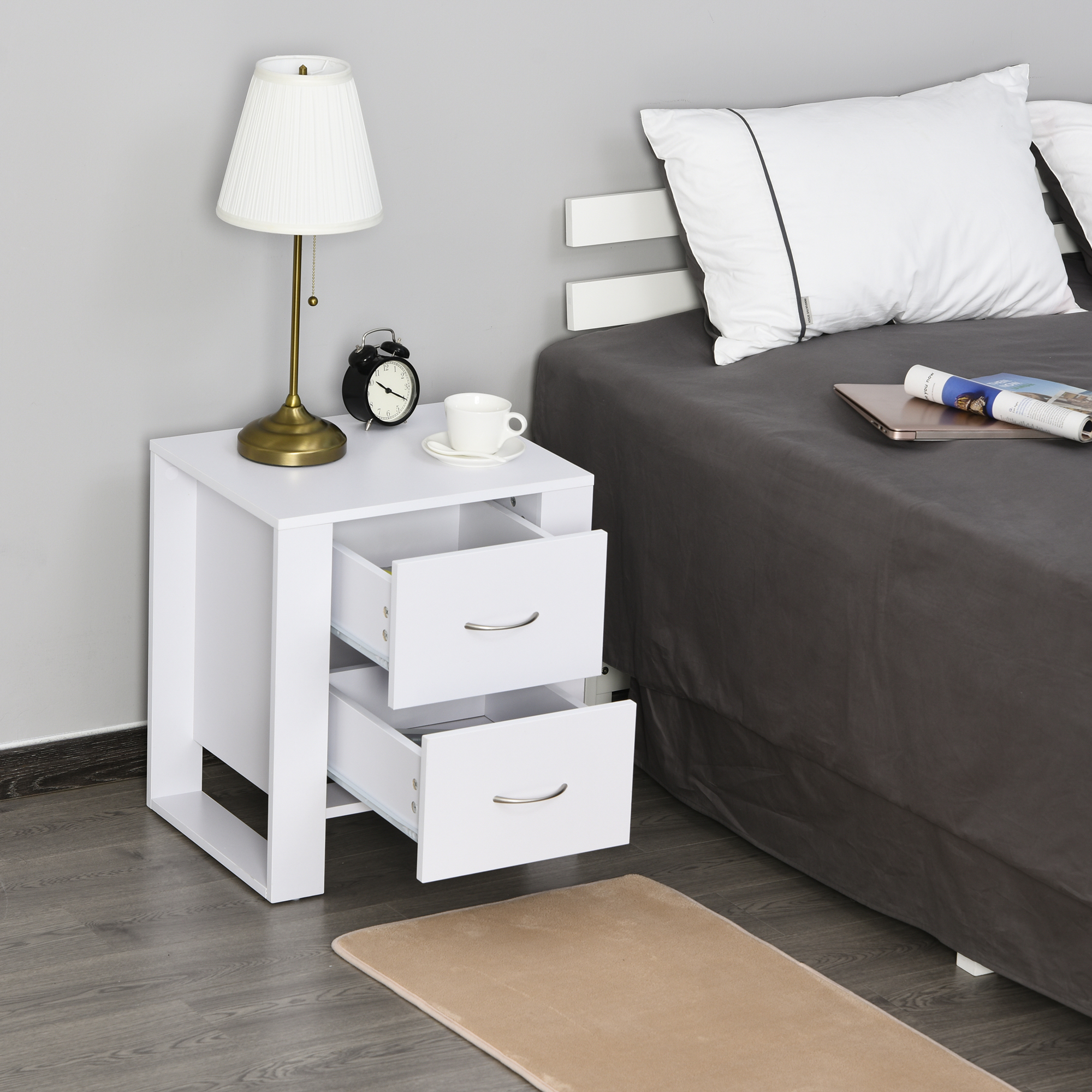 Una mesita de noche blanca con dos cajones está al lado de una cama con ropa de cama oscura.