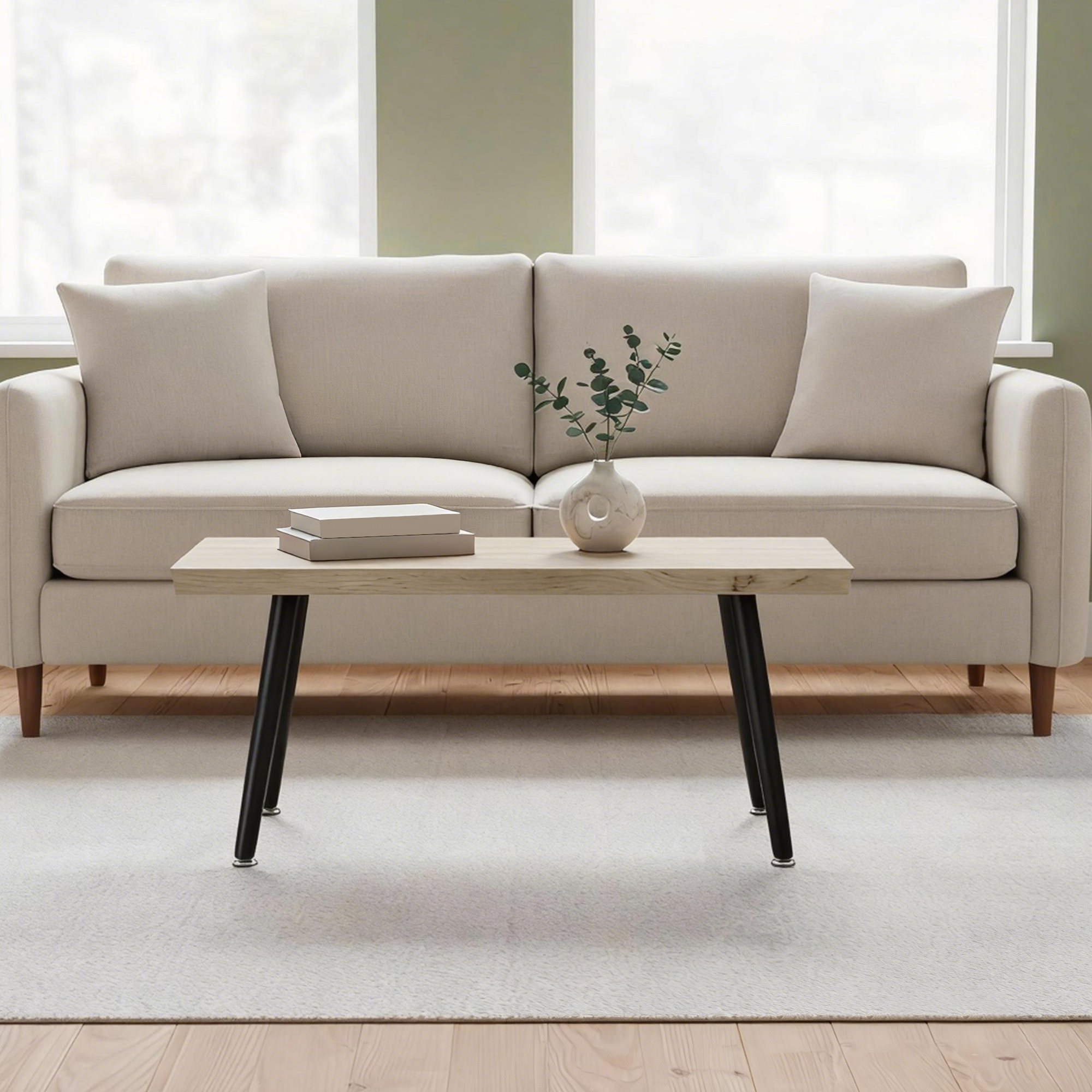 Un sofá beige con cojines está frente a una ventana. Una mesa de madera con jarrón y libros está delante.
