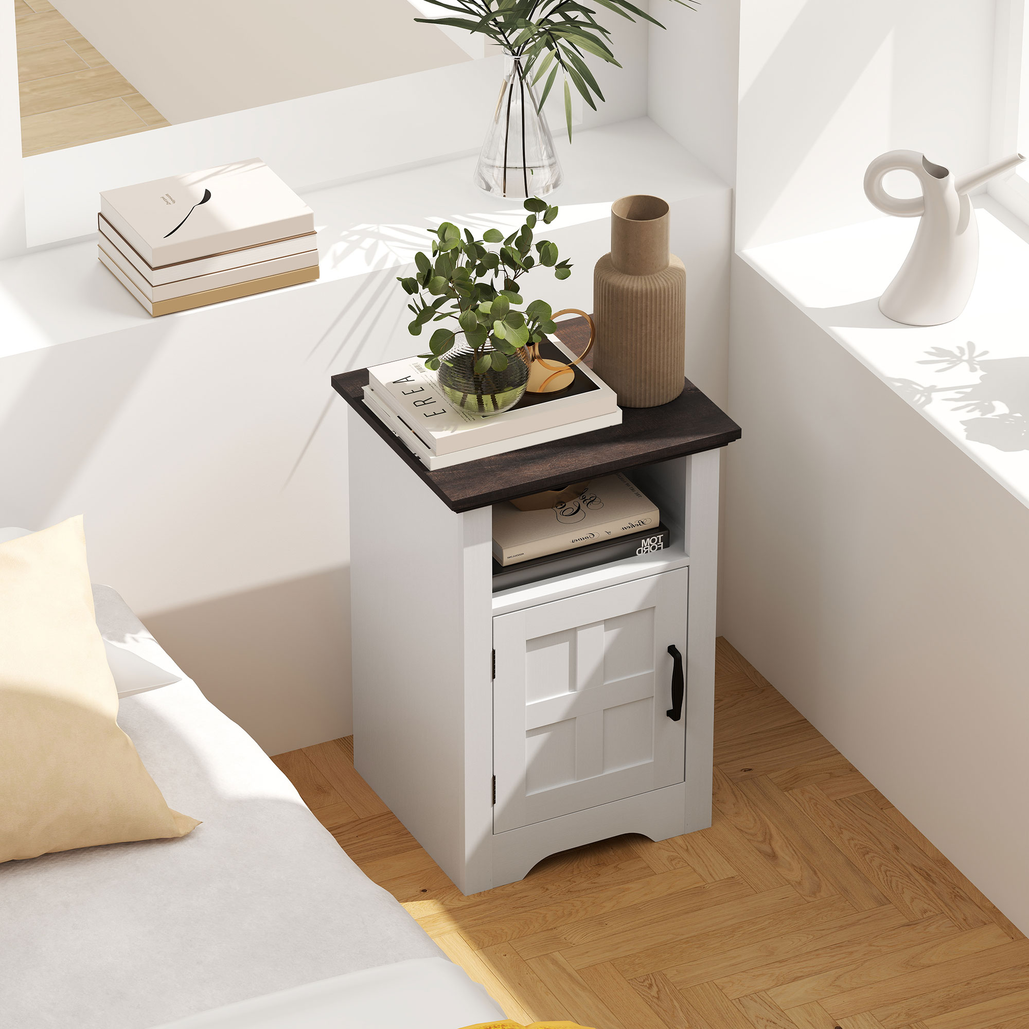 Una mesita de noche blanca con tapa oscura, con libros y plantas, junto a una cama.