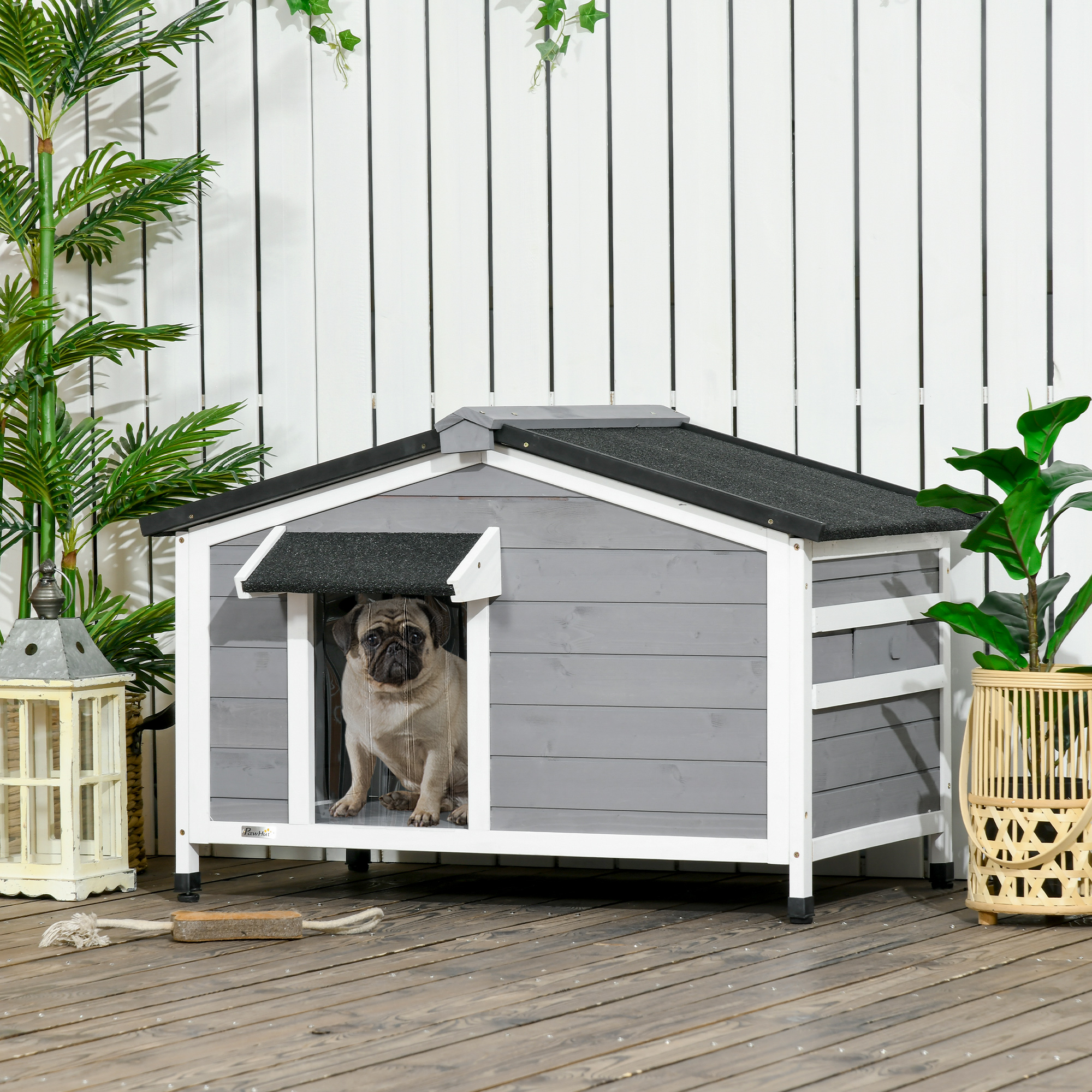 Un pug se sienta dentro de una casa para perros gris con ribete blanco contra una valla de madera.