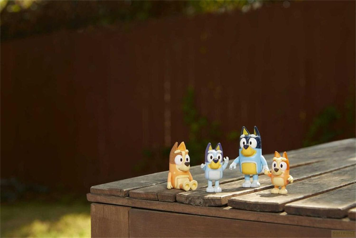 Quatre figurines Bluey se tiennent sur une surface en bois devant une clôture.
