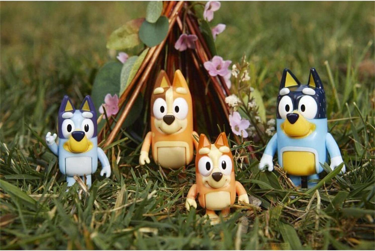 Quatre jouets Bluey dans l'herbe devant un petit tipi fait de branches et fleurs.