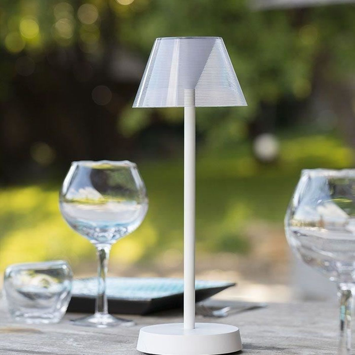 Eine weiße Tischlampe steht auf einem Holztisch mit Gläsern im Freien.