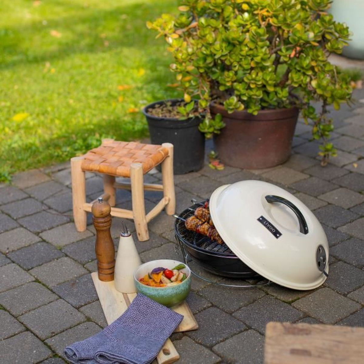 Ein Grill mit Fleischspießen, ein Hocker, Gewürzstreuer und ein Gemüseschälchen auf einer Terrasse.