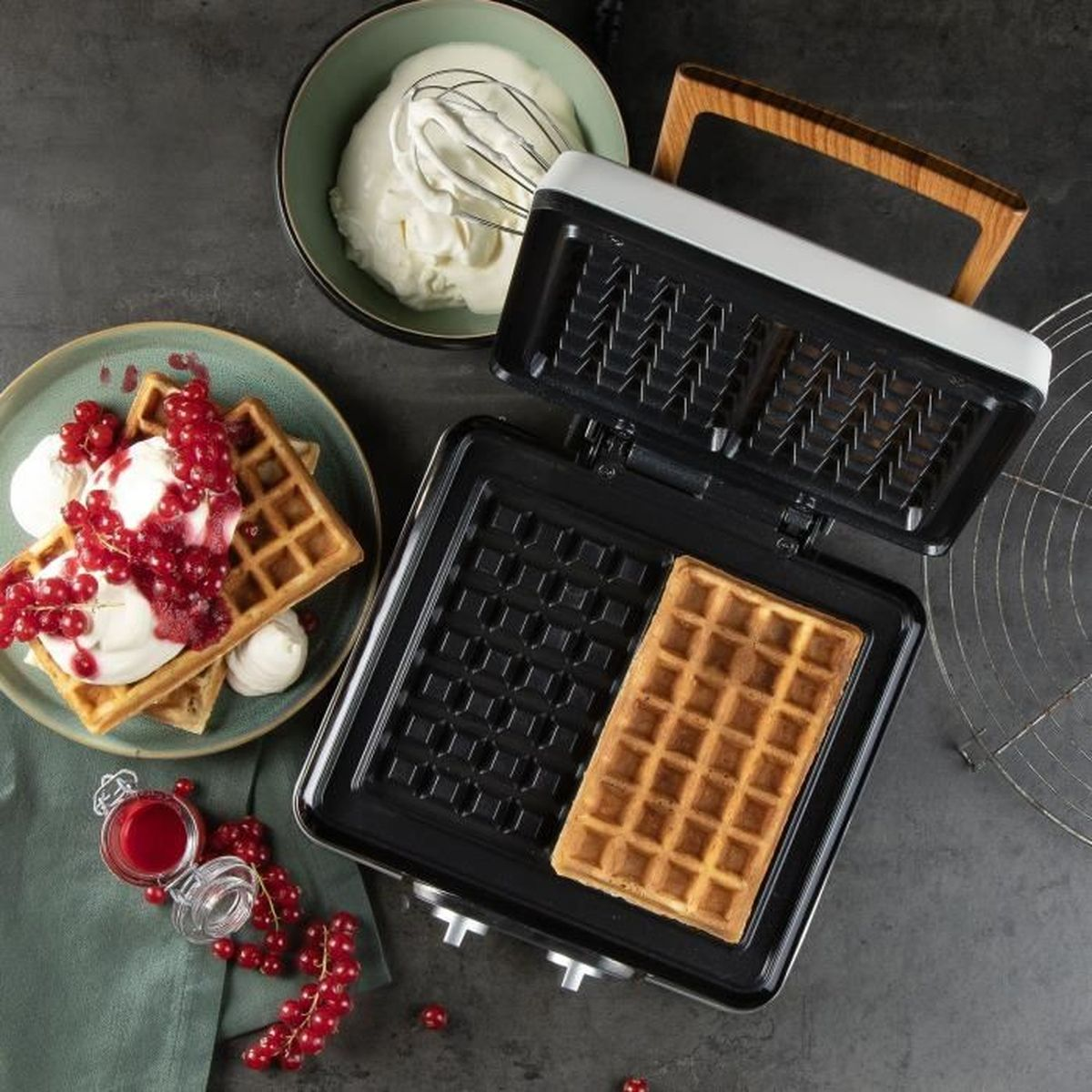 Una piastra per waffle con waffle, gelato, bacche e panna. Un waffle è in cottura.