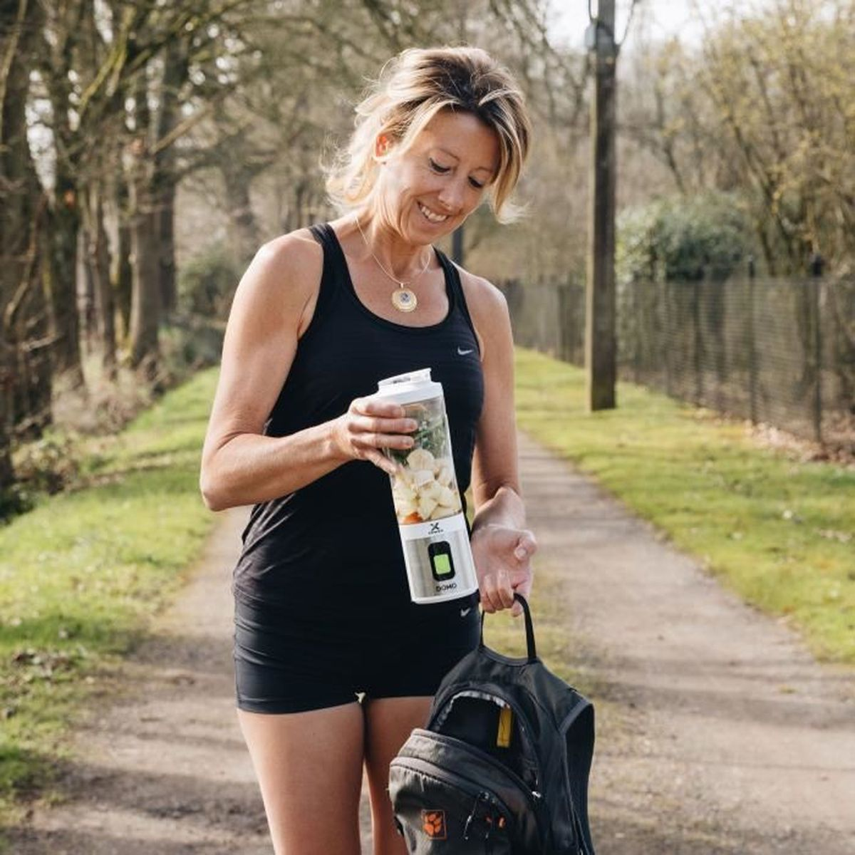 Frau in schwarzer Sportkleidung hält tragbaren Mixer mit Obst, geht auf Weg.