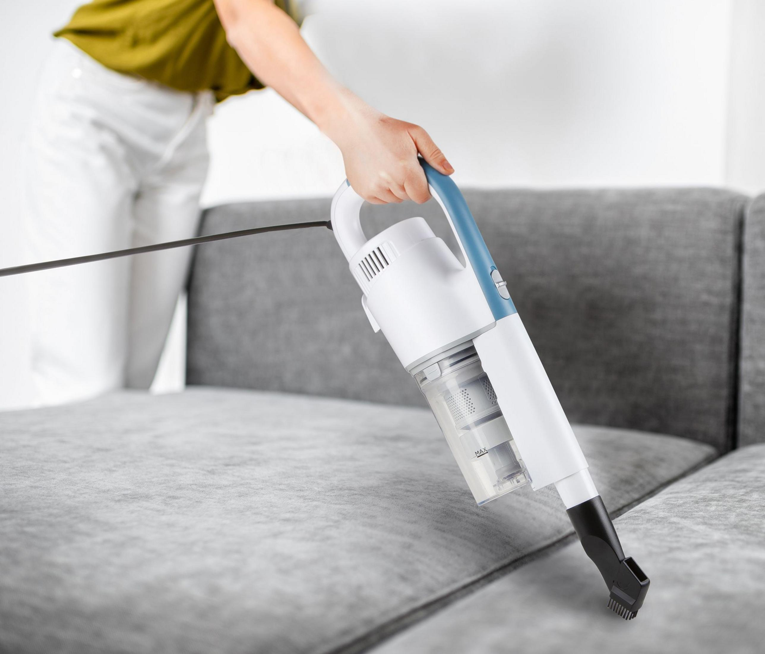 Eine Hand hält einen weißen und blauen Handstaubsauger, um ein Sofa zu reinigen.