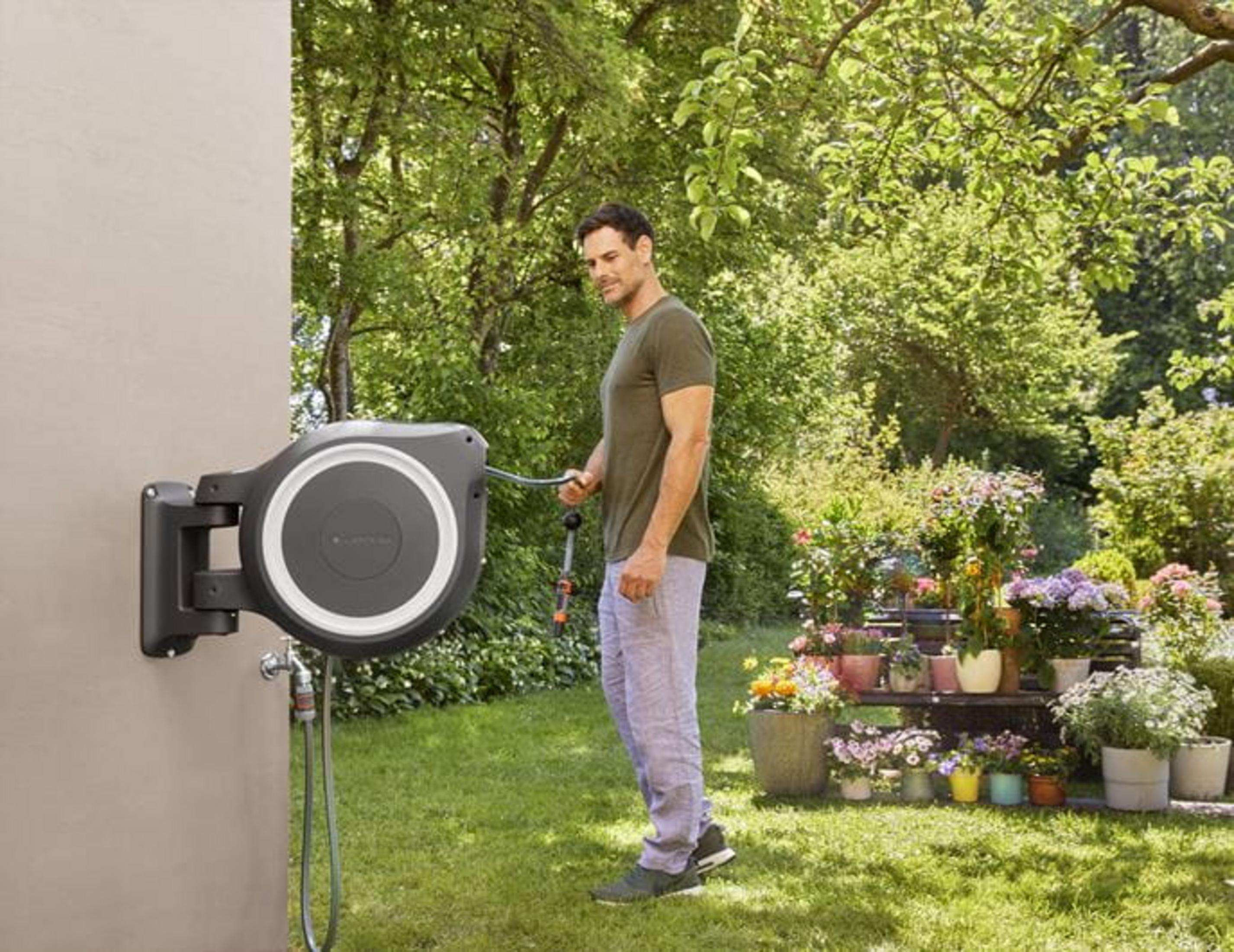 Ein Mann mit Gartenschlauch vor einem Wand-Schlauchaufroller und blühenden Pflanzen.