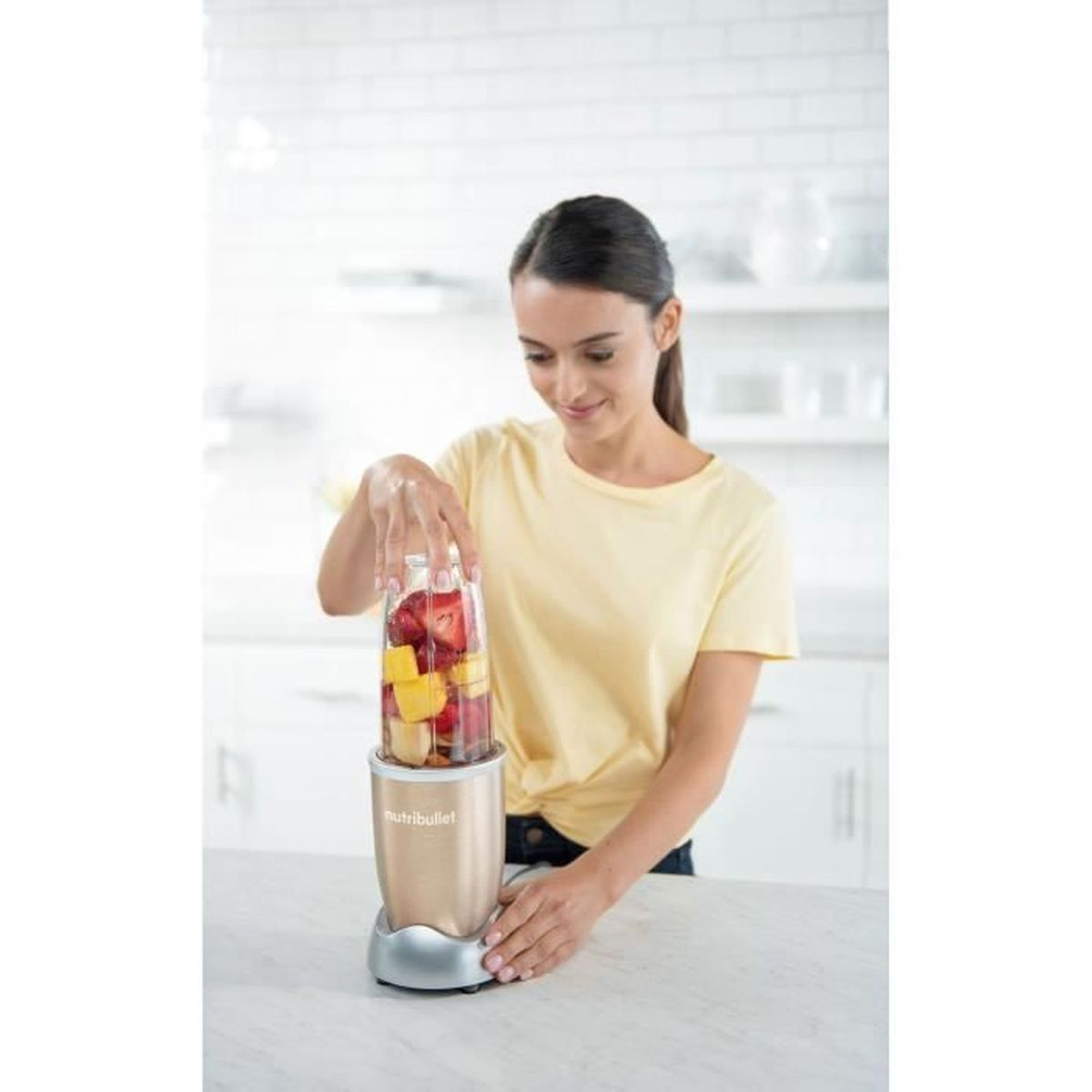 Una donna in maglietta gialla mette una tazza di frutta in un frullatore.