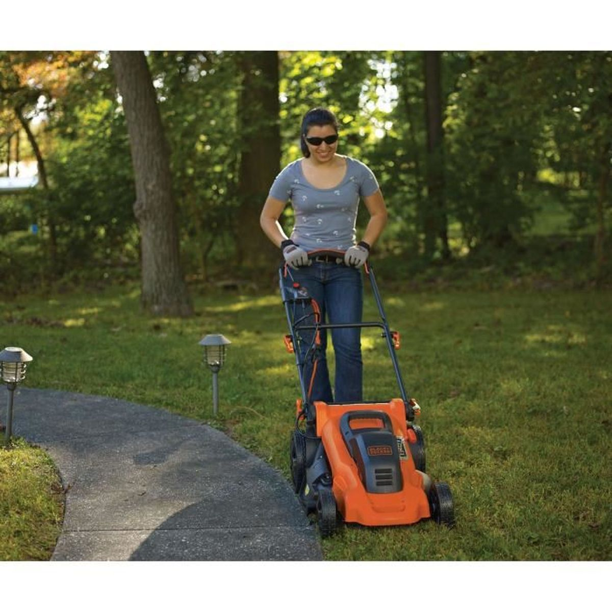 Eine Frau mäht mit einem orangefarbenen Rasenmäher auf einem Rasen neben einem Weg.