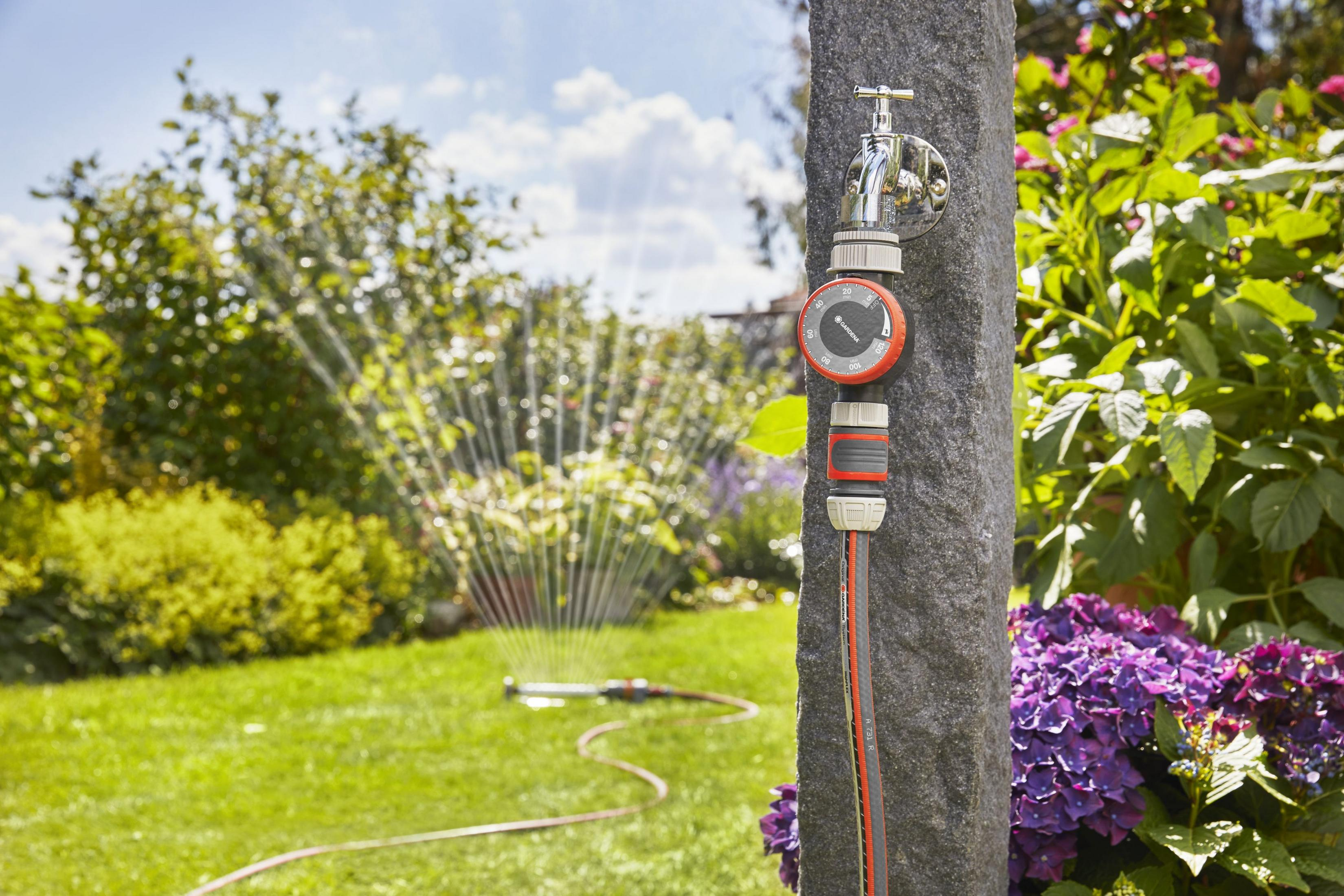 Wasserhahn mit Zeitschaltuhr an Steinsäule, im Garten wird Rasen gesprengt.