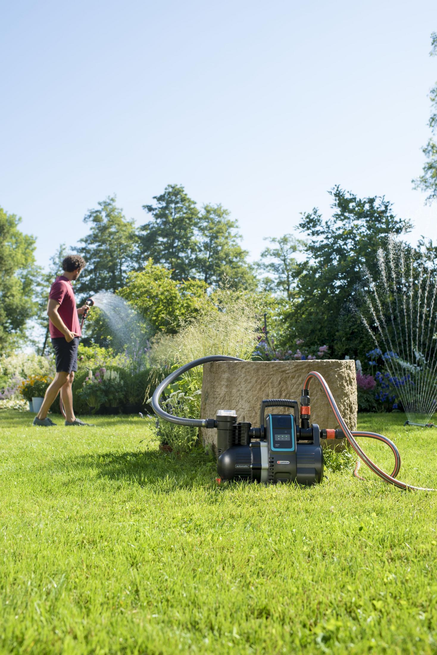 Mann bewässert Garten; Gartenpumpe auf Rasen mit Bewässerungsanlage im Hintergrund.