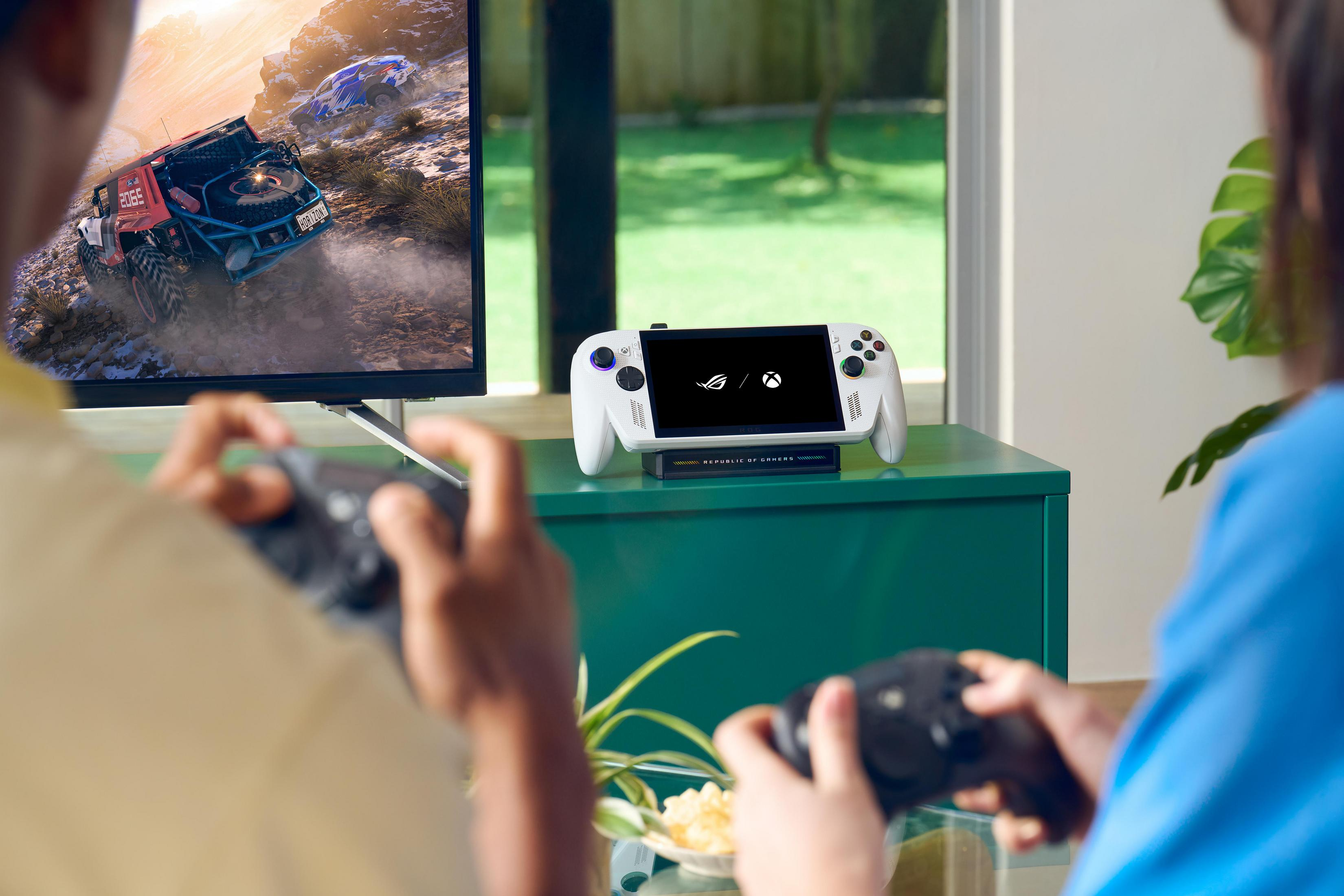 Zwei Personen spielen ein Rennspiel auf einem Fernseher und halten Controller. Ein Handheld-Gerät steht daneben.