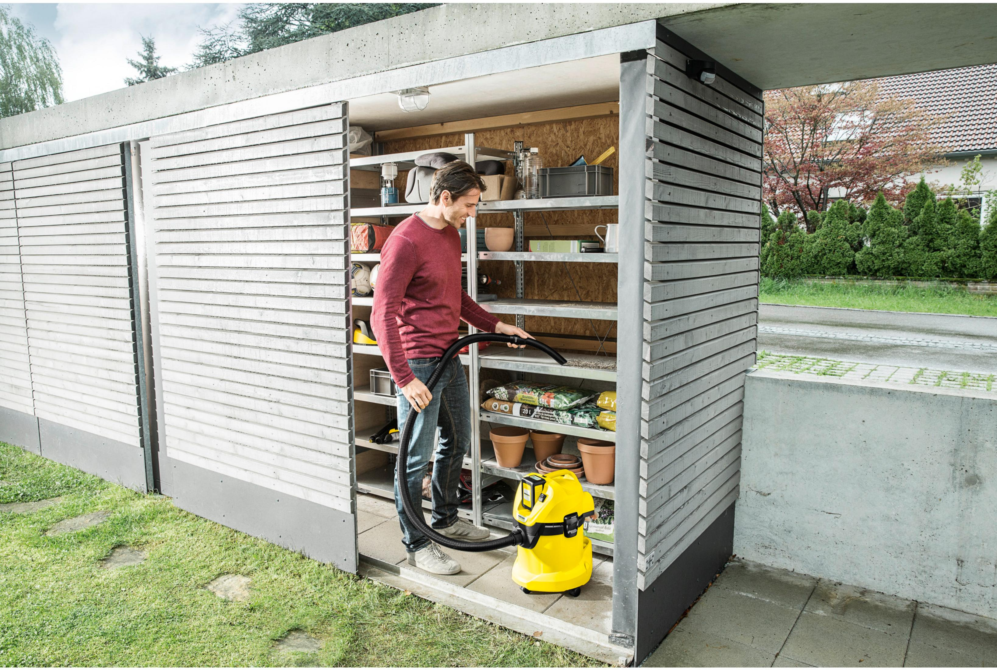 Homme passe l'aspirateur dans un abri de jardin. Étagères remplies.