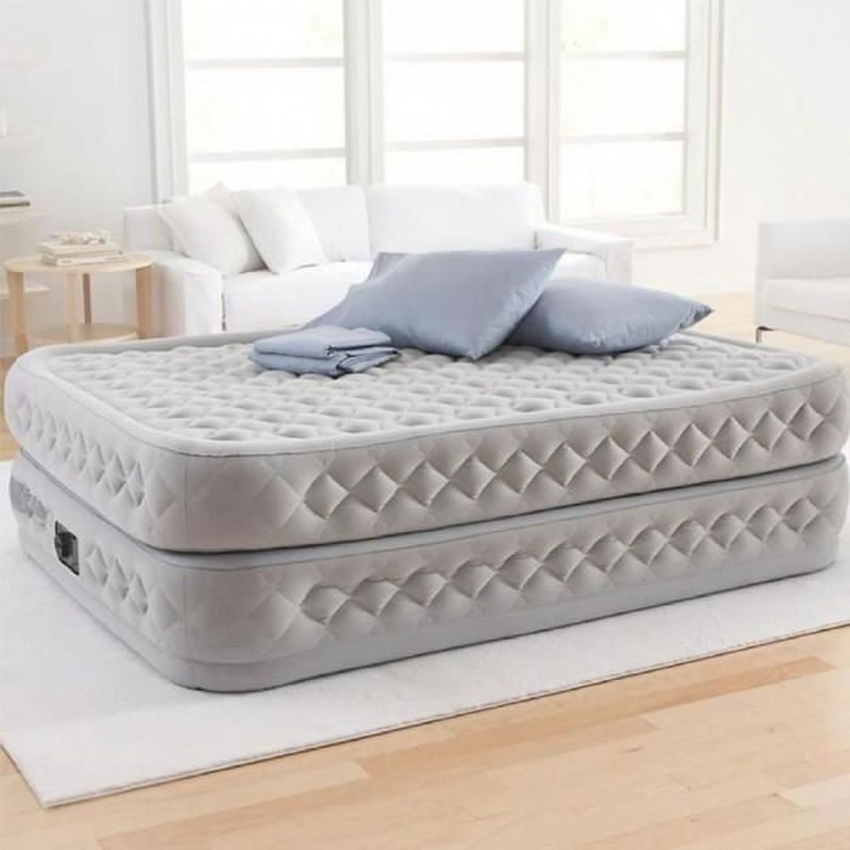 Colchón de aire gris de dos niveles con almohadas y ropa de cama azules. Fondo de sala.