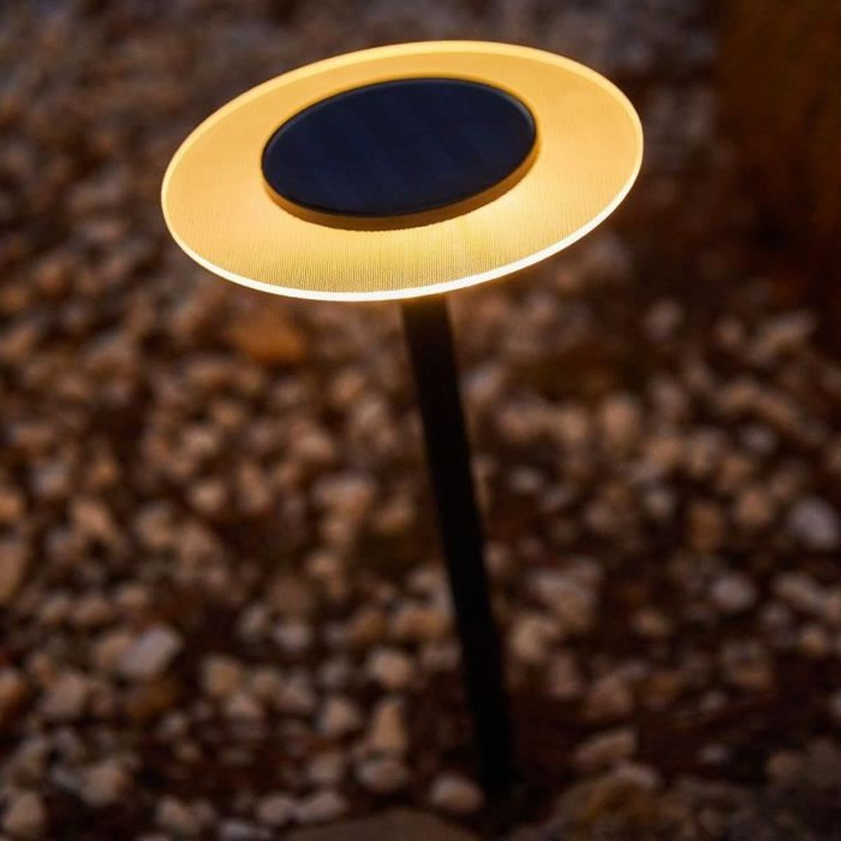 Ein kreisförmiger Außenleuchter mit warmem Licht auf einem Kiesweg.