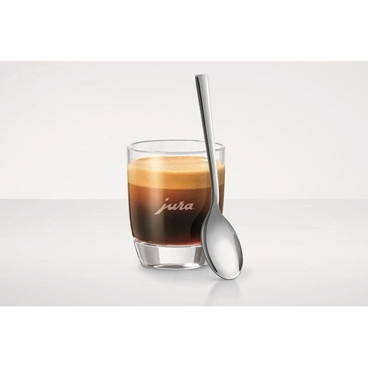 Un verre d'espresso avec une mousse crémeuse et une cuillère à côté sur fond blanc.